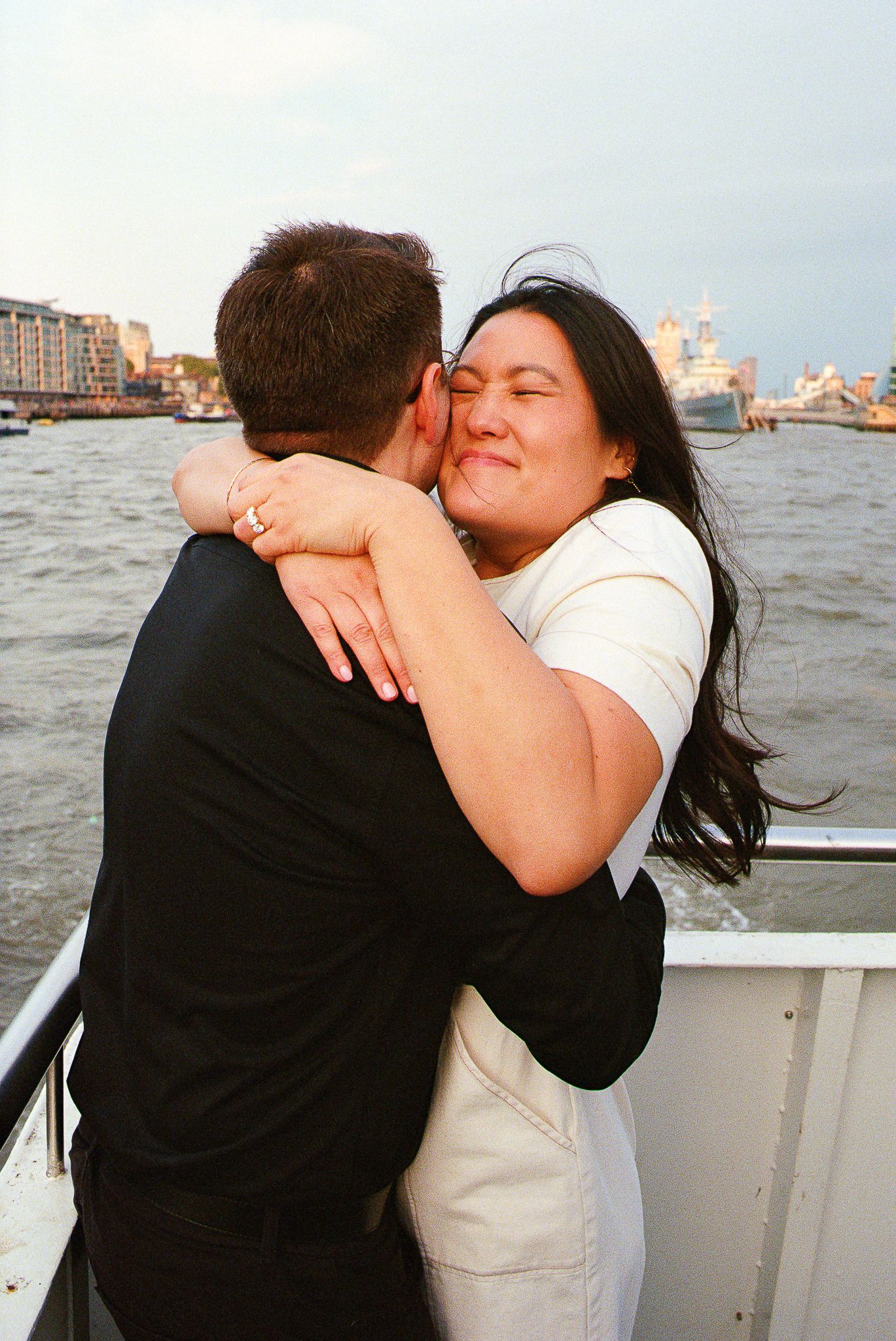 Two people share an intimate embrace on a boat with London's skyline in the background.