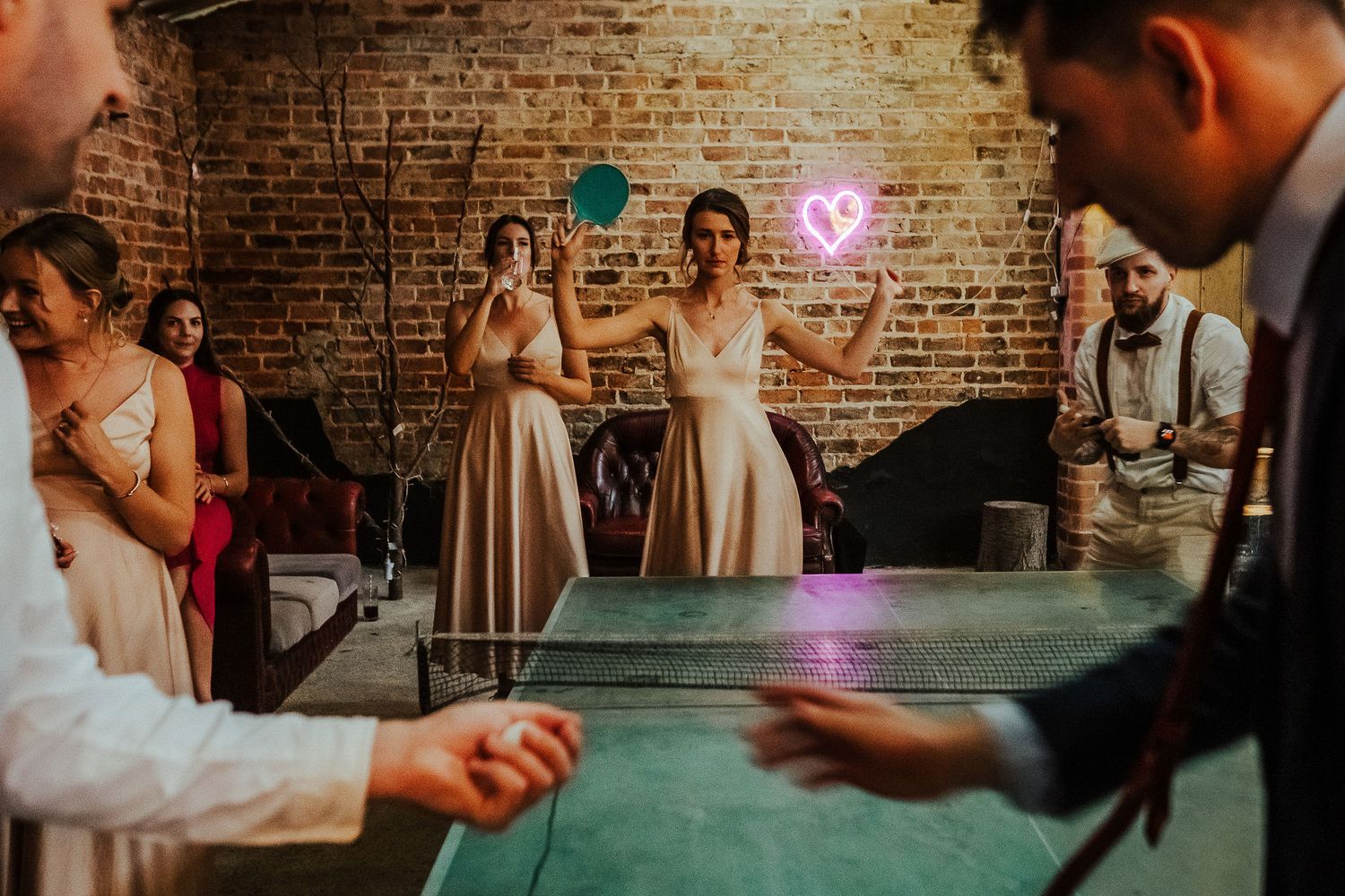 Wedding guests playing ping pong at a reception with a neon heart sign in background.