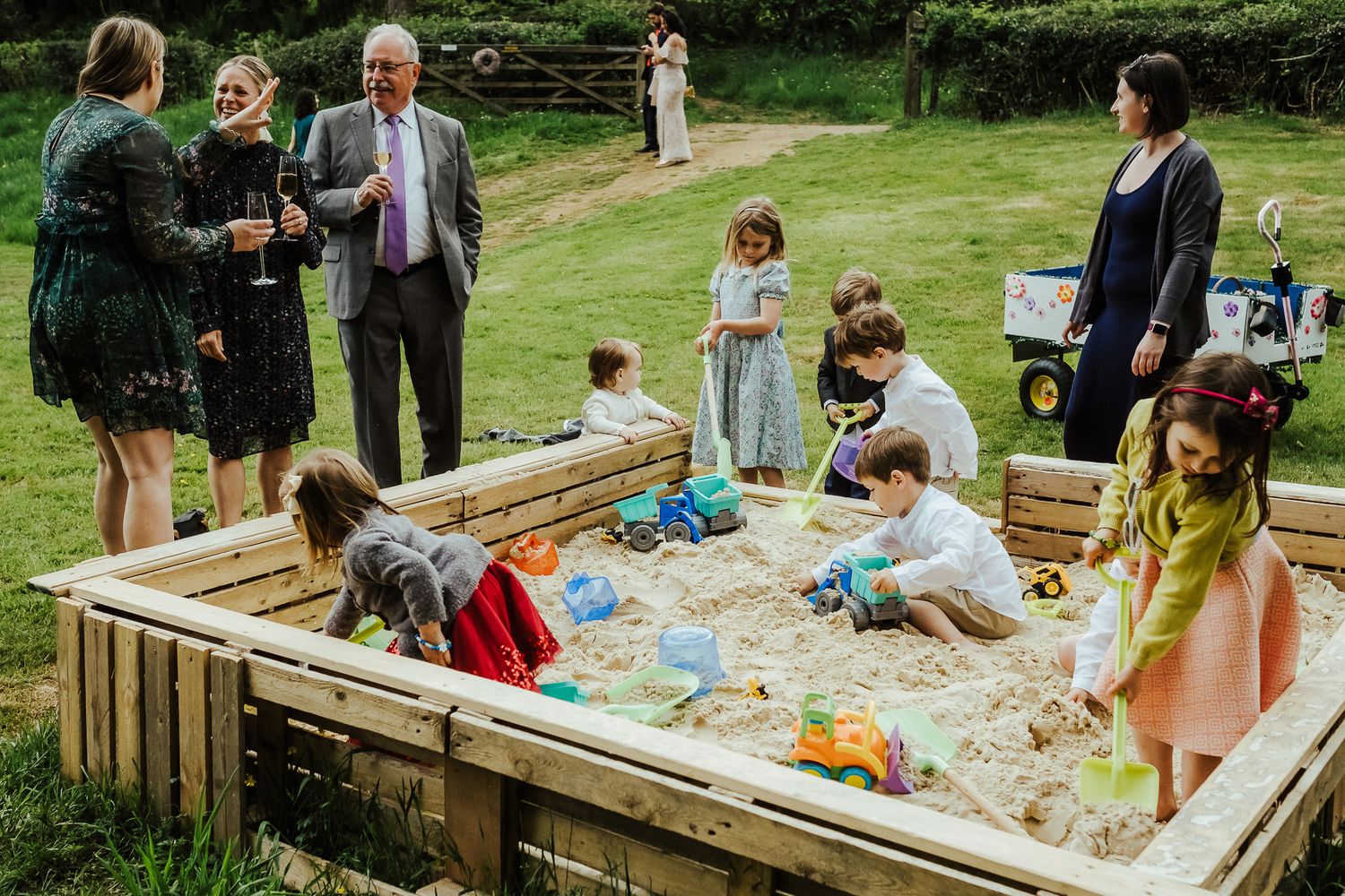 Children play in a large sandbox while adults watch during an outdoor wedding reception.