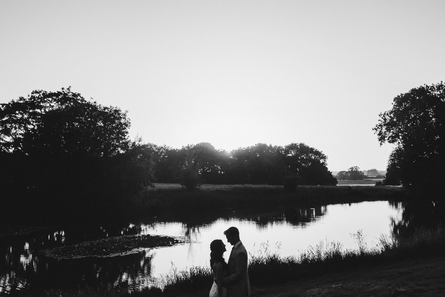 Silhouettes of a couple standing by a serene lake at sunset creating a romantic black and white scene.