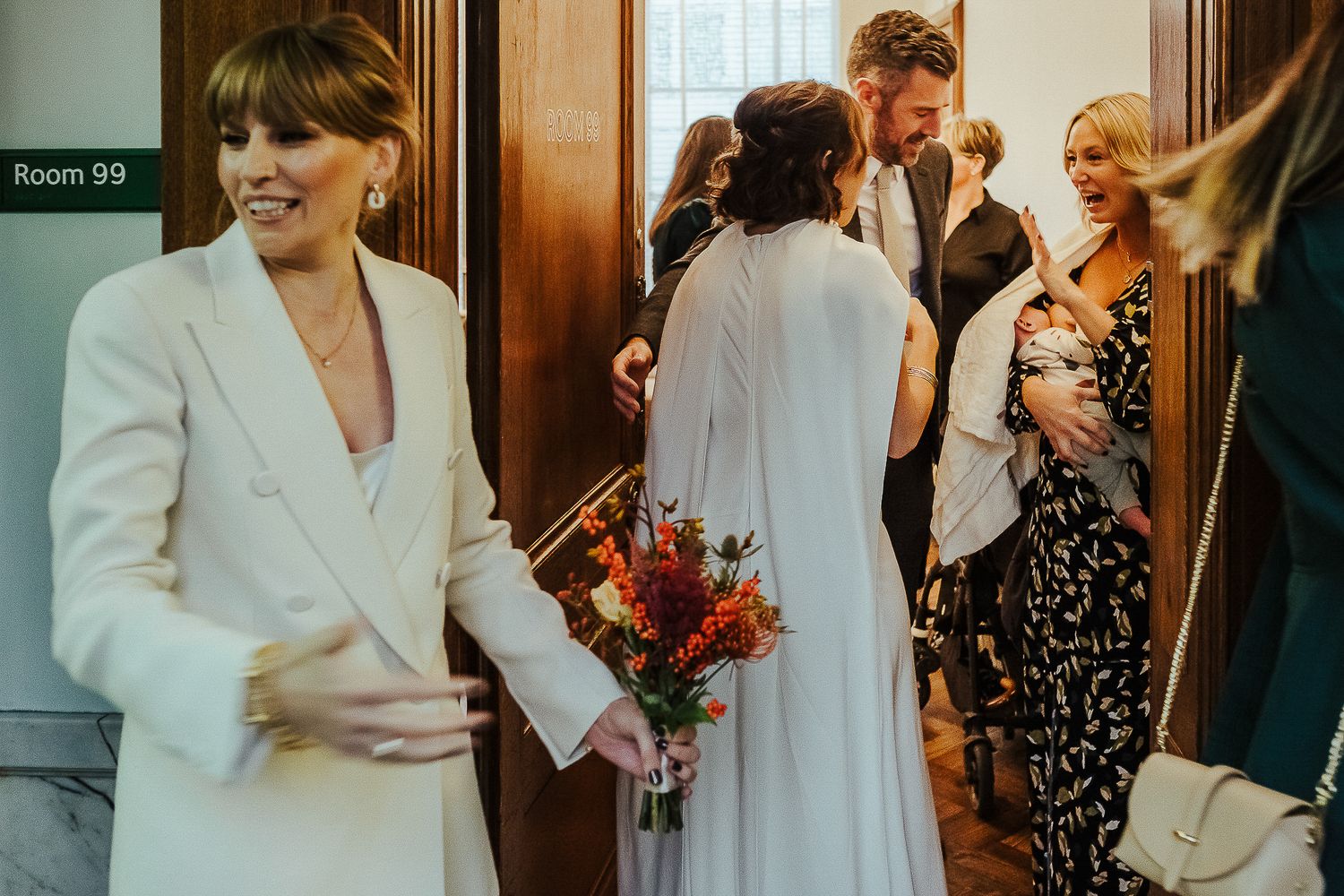 A bride in white carries a vibrant bouquet while entering through a doorway with guests.