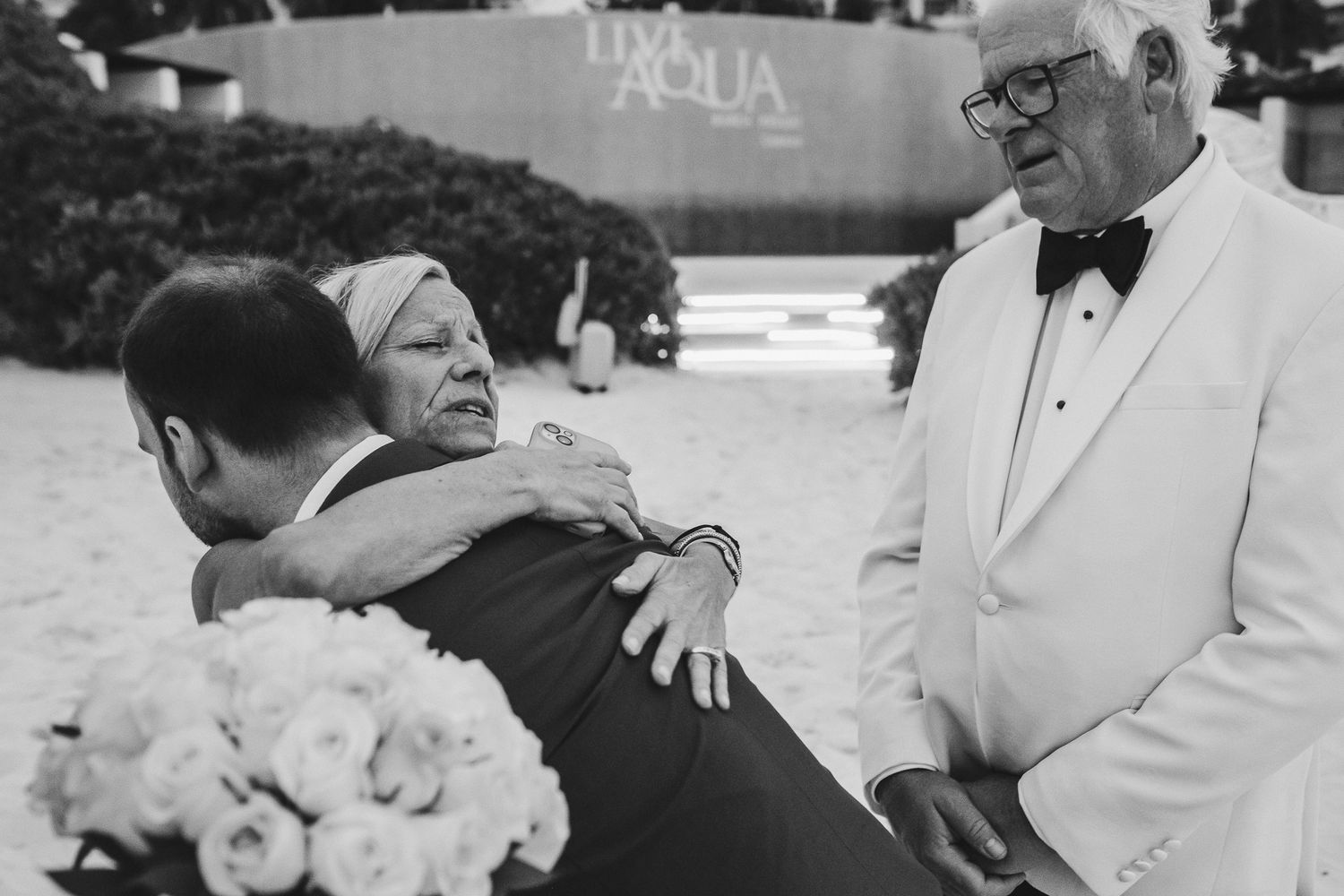 Black and white candid photo of emotional wedding reception moment with guests embracing.