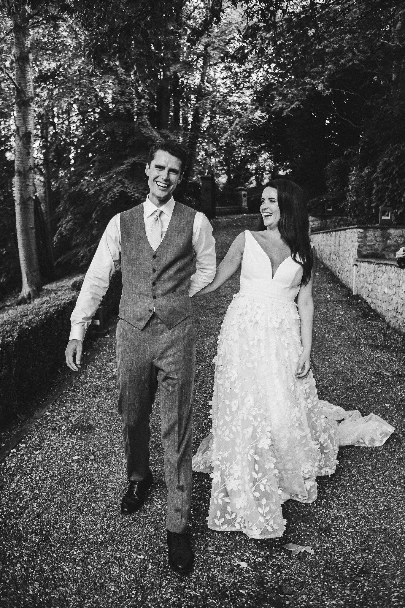 Bride and groom walk hand in hand along tree-lined path in black and white.