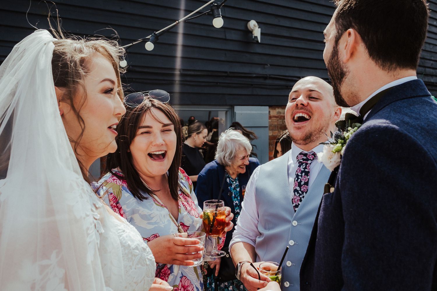 Wedding guests share laughter and drinks during outdoor reception celebrations.