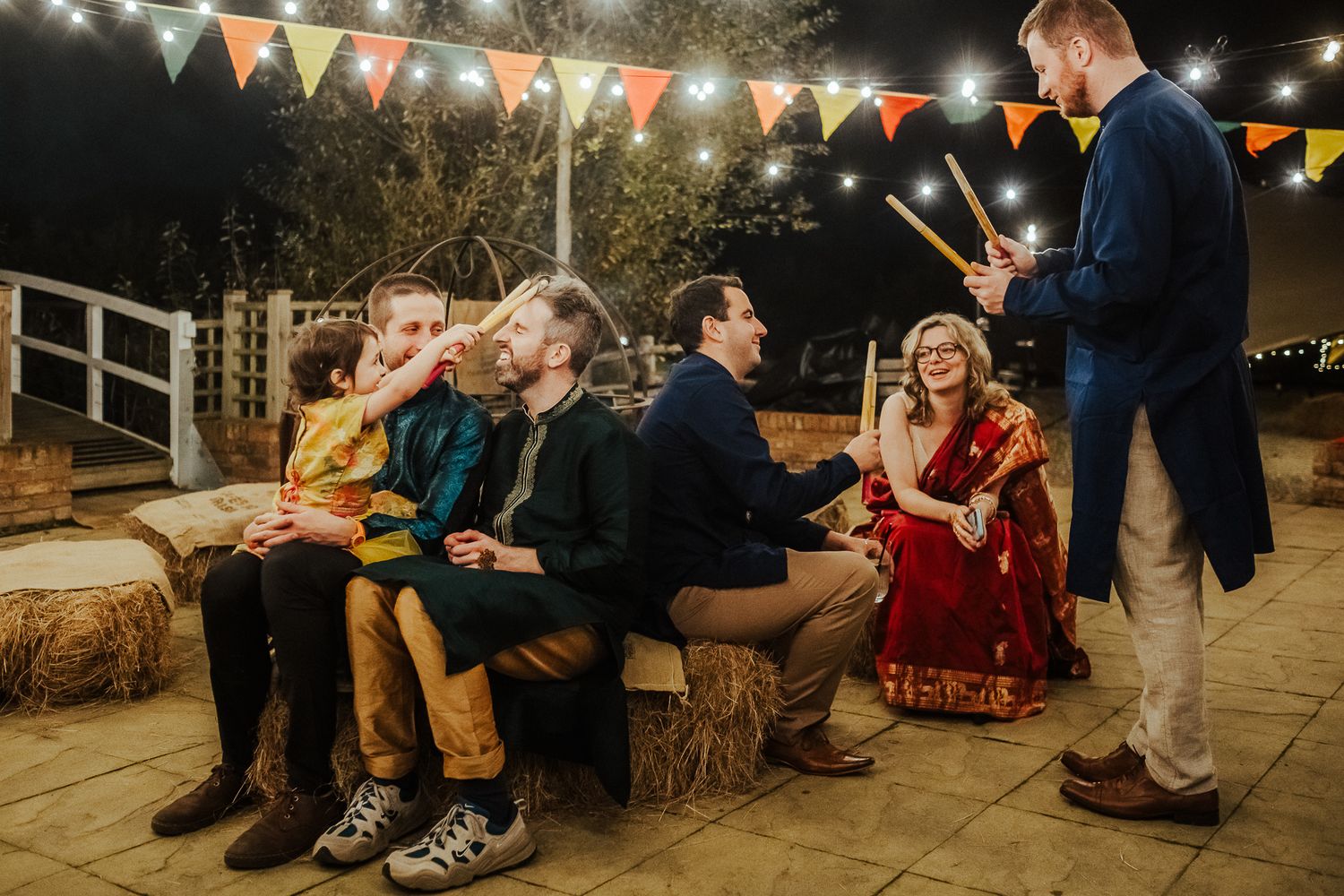 Casual outdoor gathering with guests seated on hay bales under festive bunting at night.