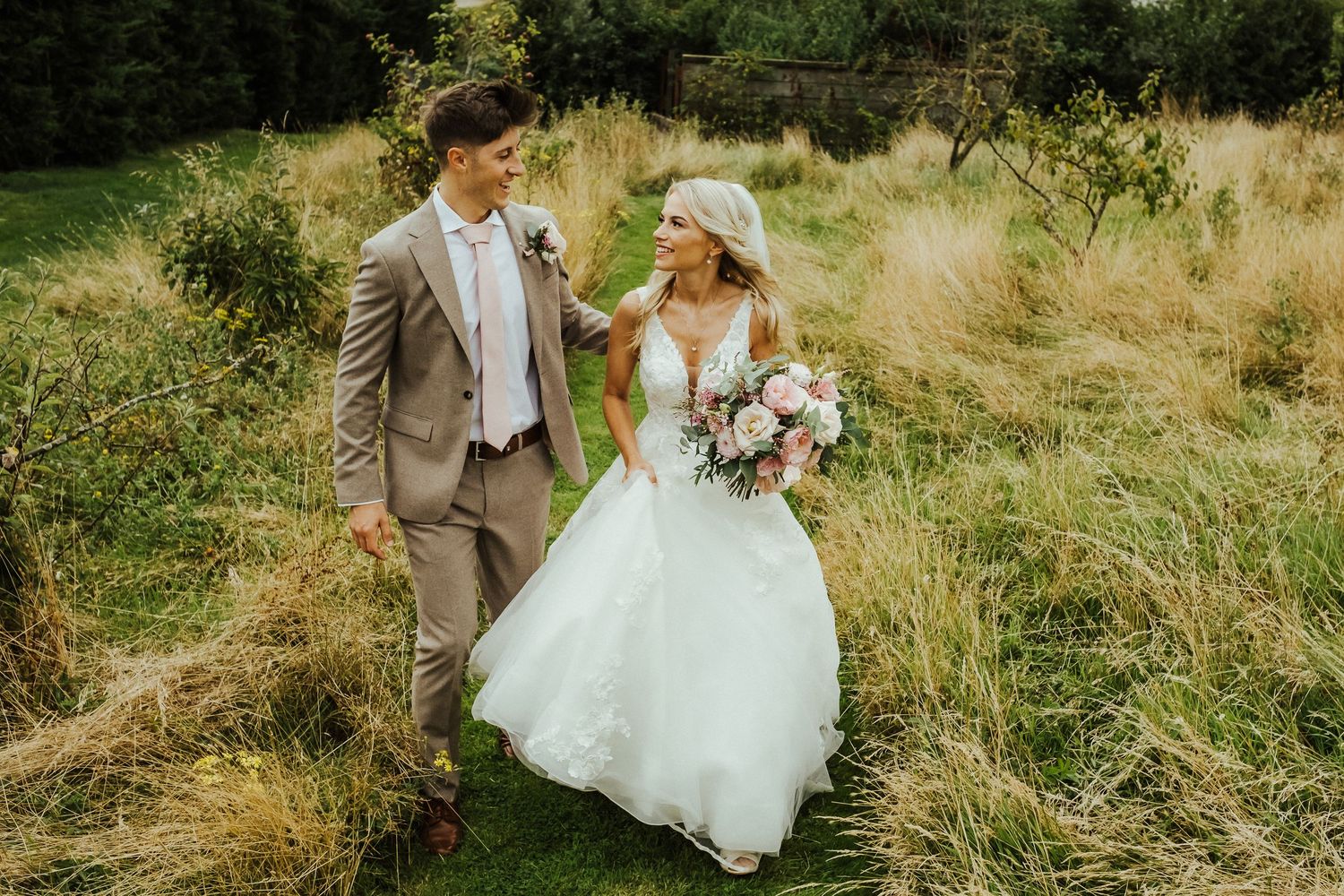Newlyweds share a laugh while walking through tall summer grass.