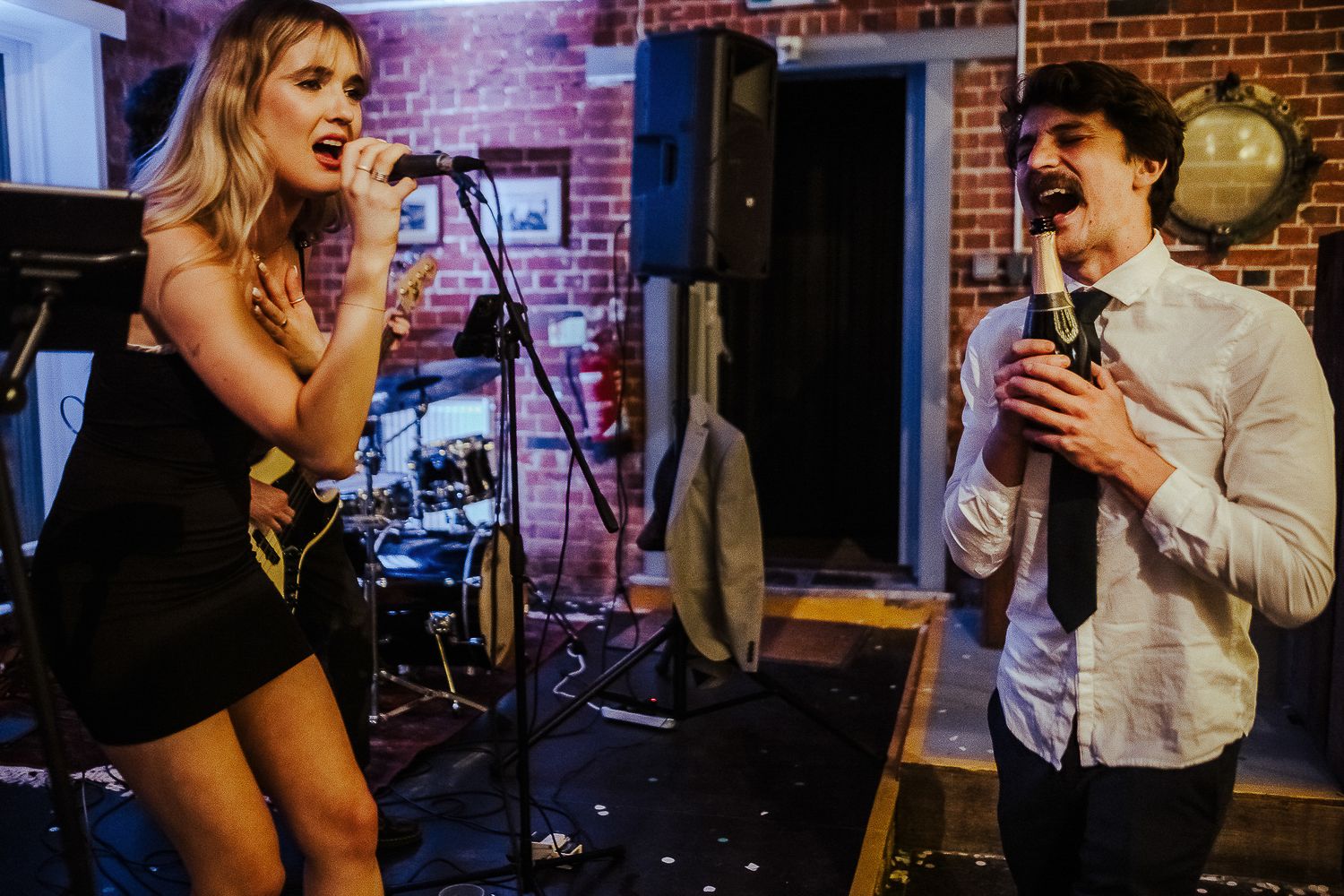 Two people performing karaoke enthusiastically in front of brick wall at party venue.