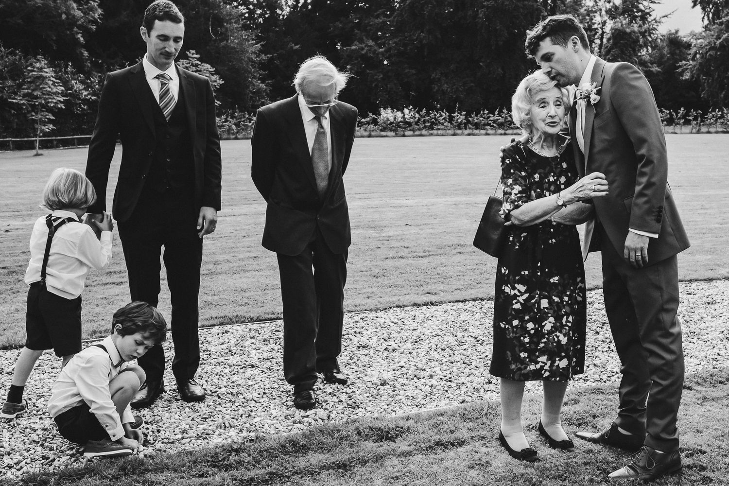 Black and white candid photo of multi-generational family members interacting at an outdoor wedding.