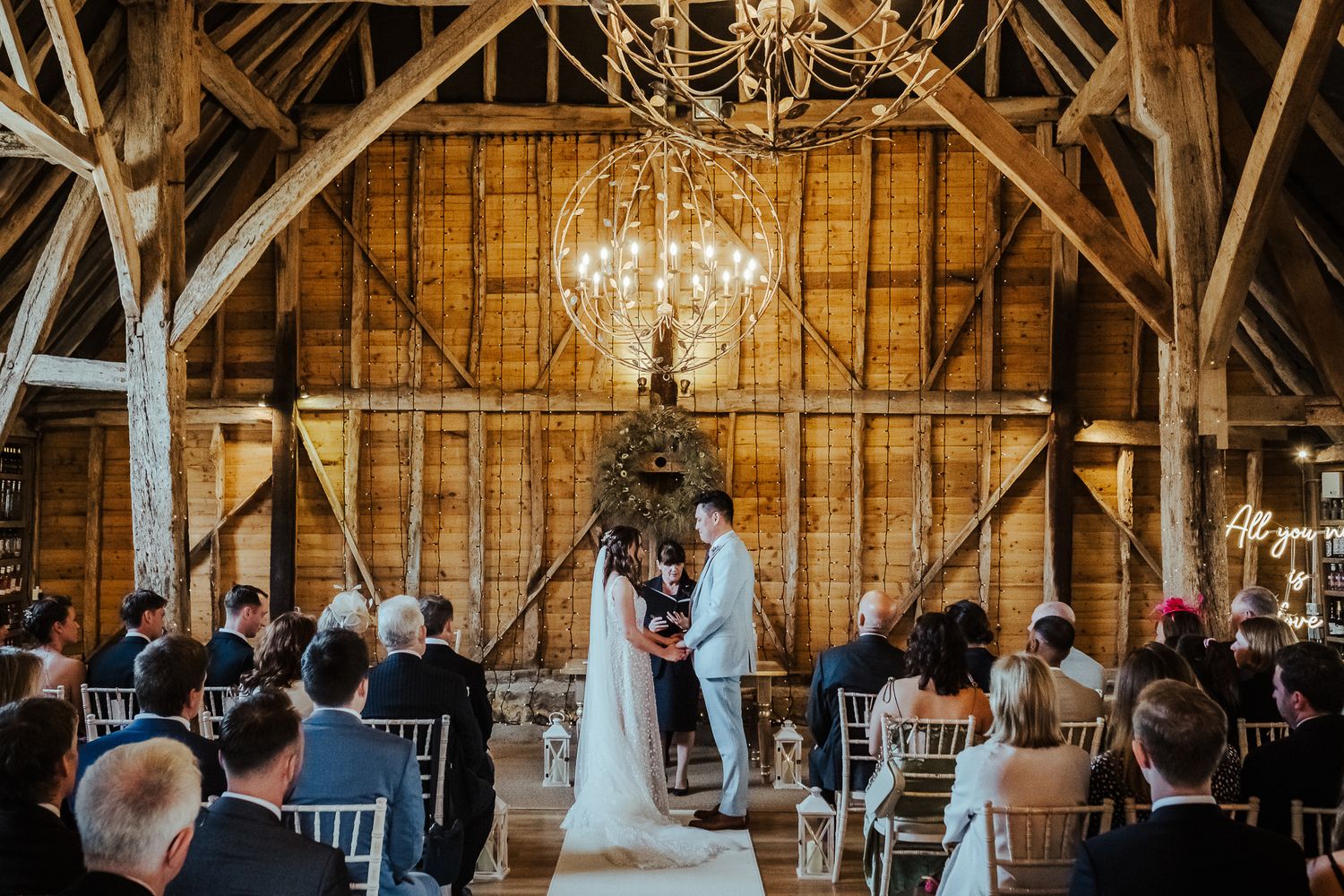 Rustic barn wedding ceremony under elegant chandelier with wooden beam backdrop.
