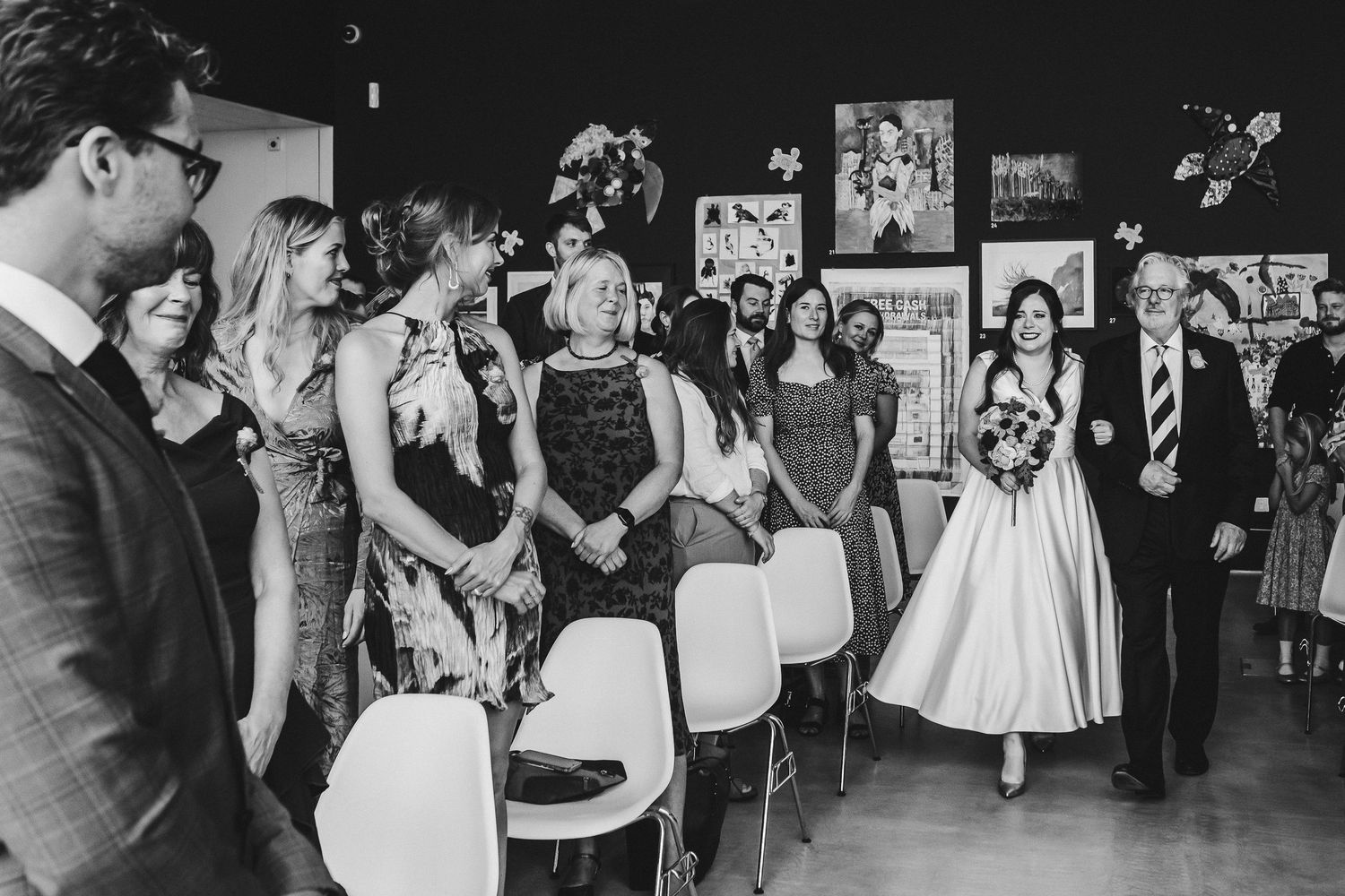 Black and white photo of guests watching wedding ceremony in modern art gallery setting.