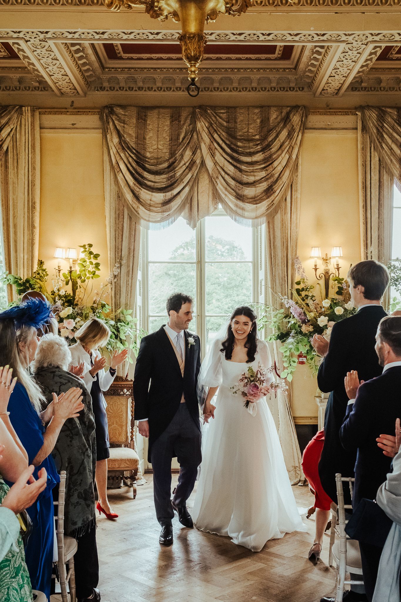 Newlyweds in an opulent ballroom with draped windows and floral decorations.