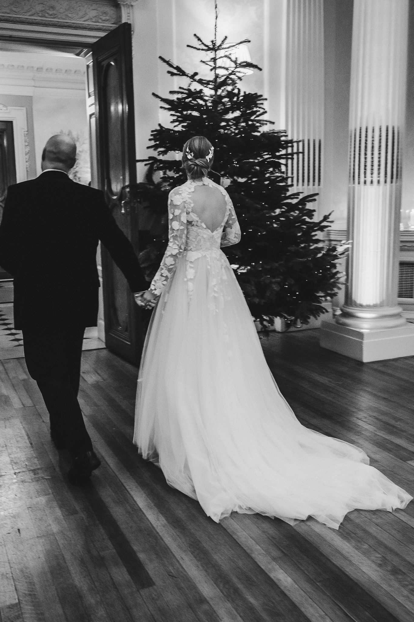 Black and white view of bride's lace-detailed dress train as couple walks past Christmas tree.