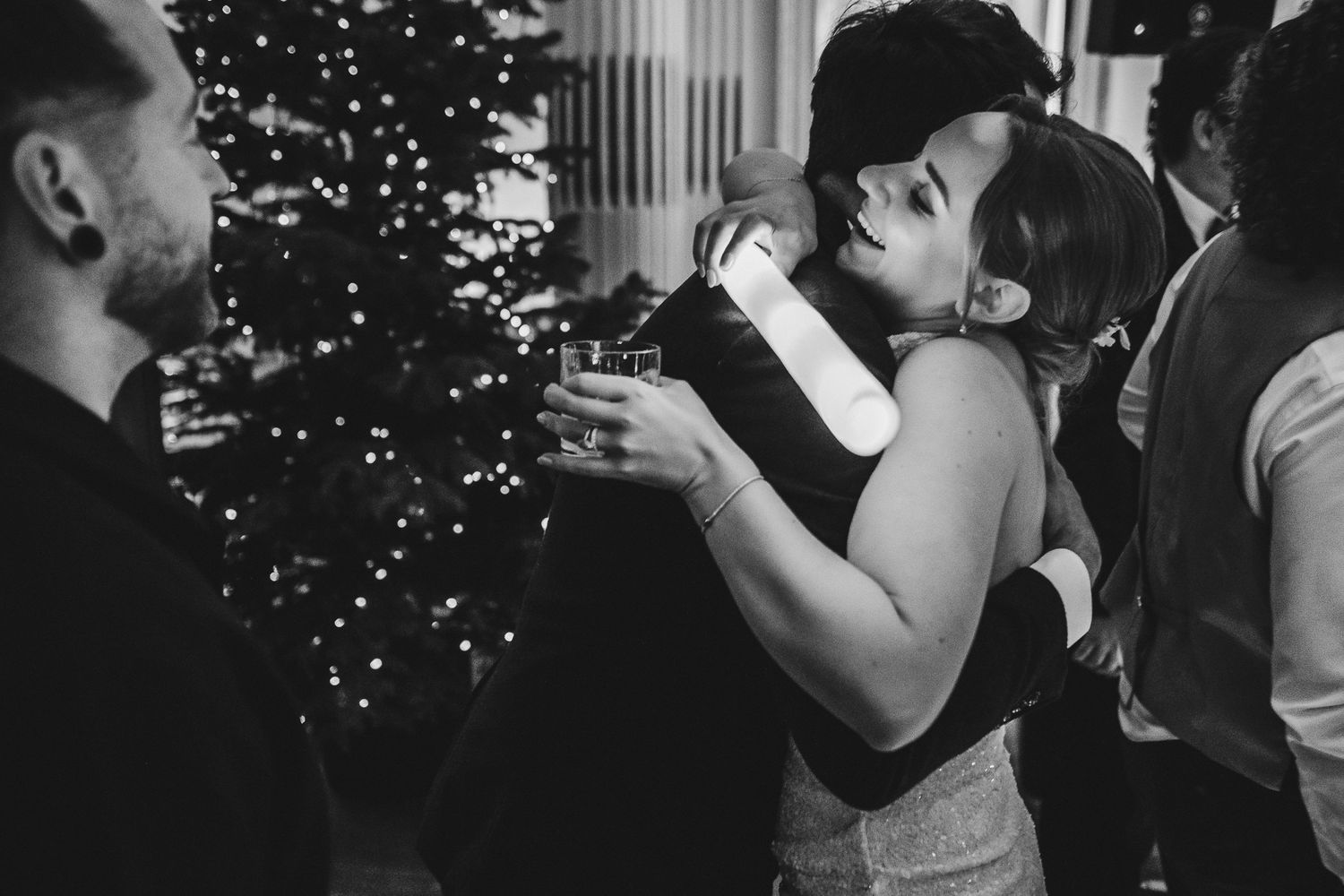 Intimate black and white photo showing guests embracing near a decorated Christmas tree at a wedding reception.