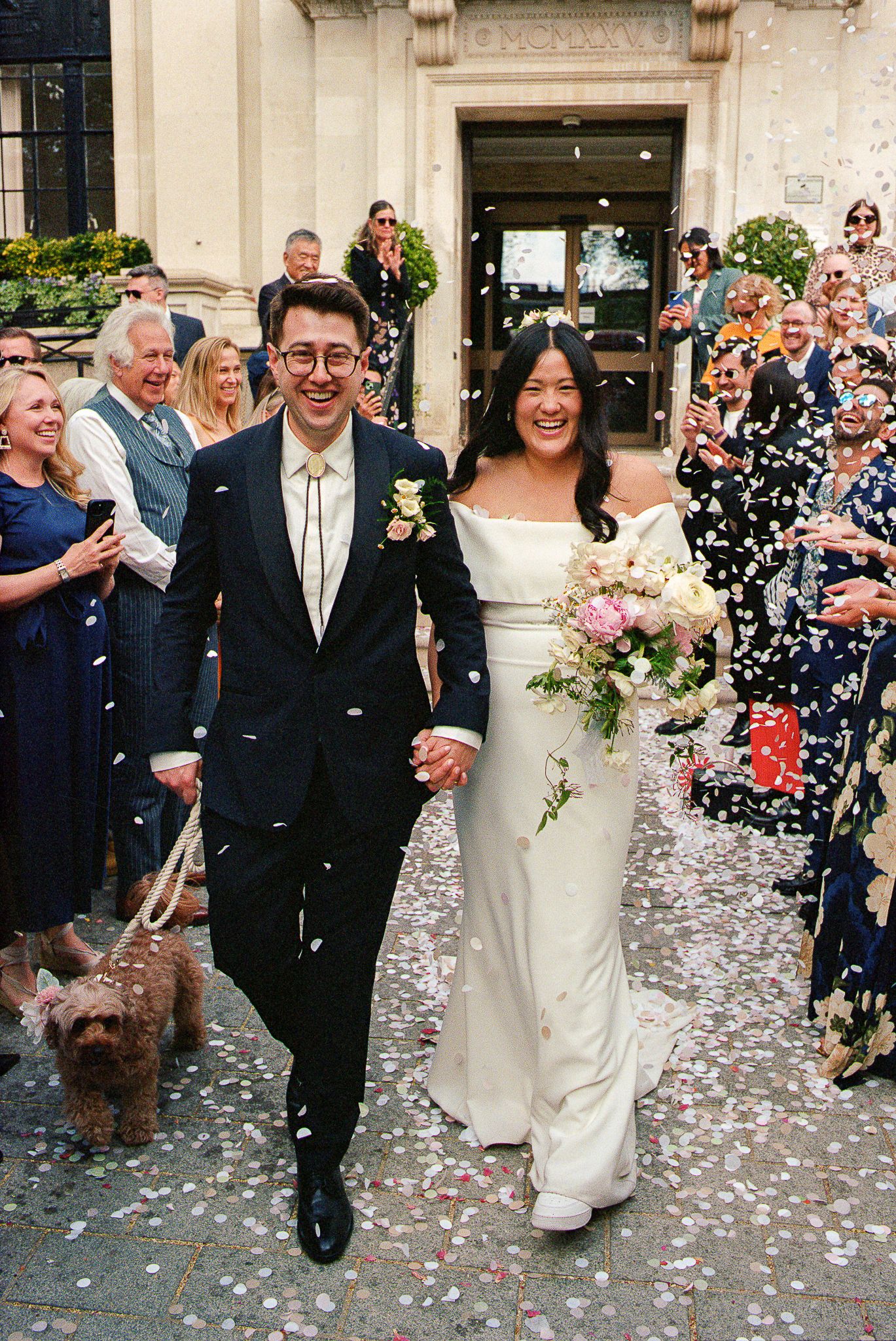 Newlyweds exit their wedding ceremony as guests shower them with confetti while their small brown dog walks alongside them.