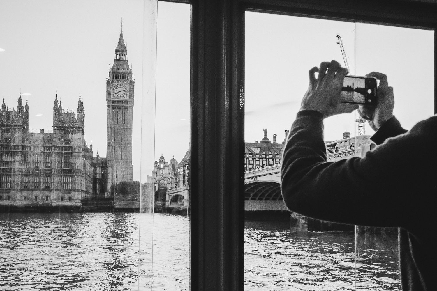 Tourist photographs iconic Big Ben and Houses of Parliament from River Thames boat in black and white London scene.
