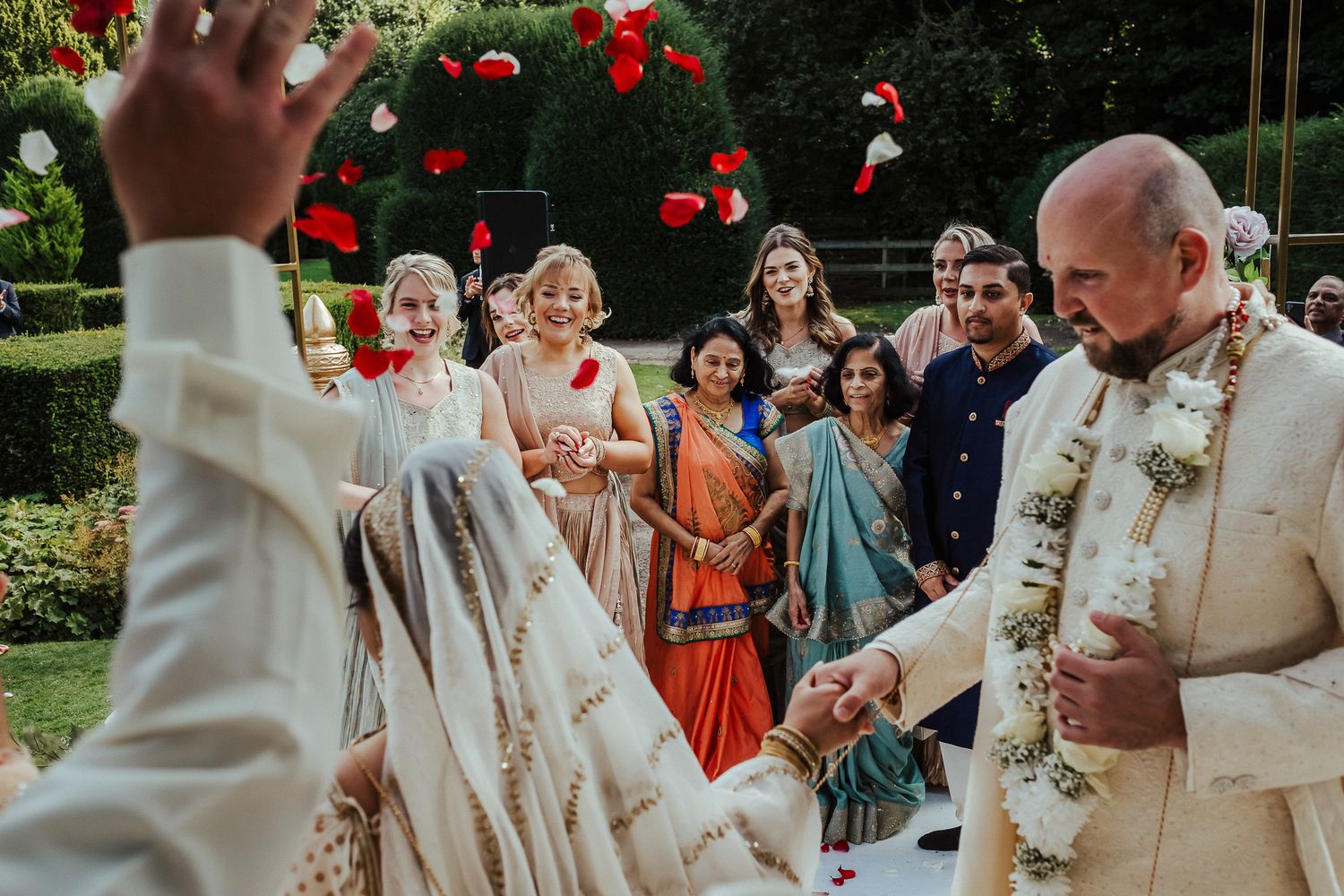 Candid outdoor wedding ceremony moment with guests in colorful attire watching ceremony in a garden setting.