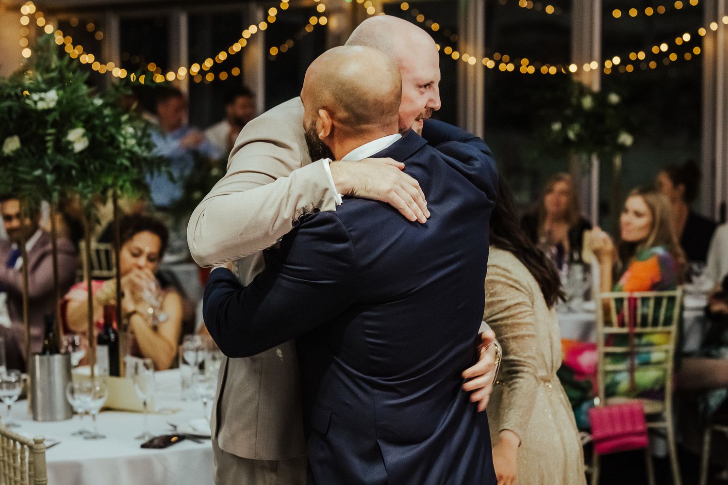 Two people share an emotional embrace at a wedding reception with string lights and table settings in the background.