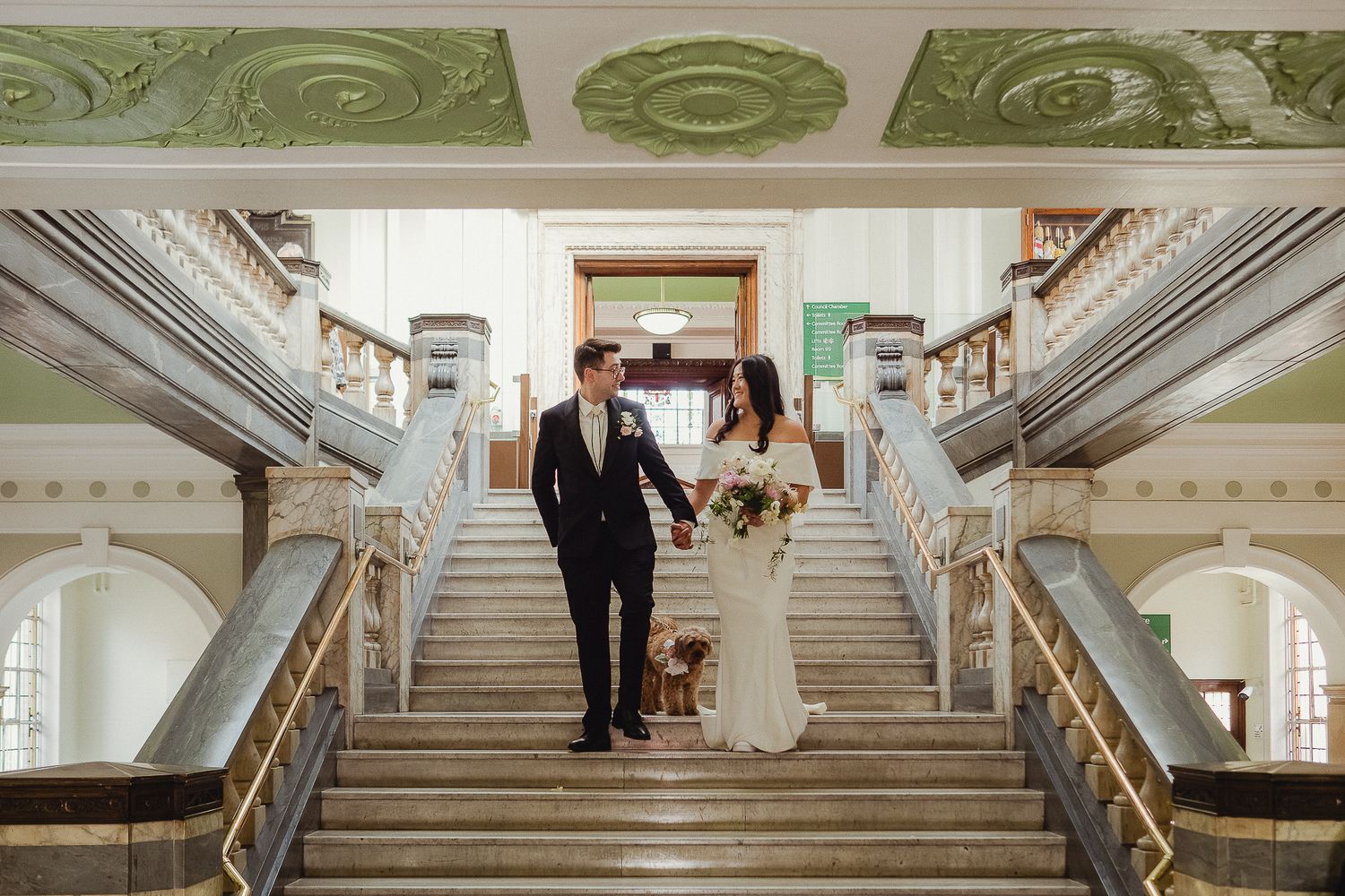 A couple descends an elegant marble staircase surrounded by ornate architectural details in a historic building.