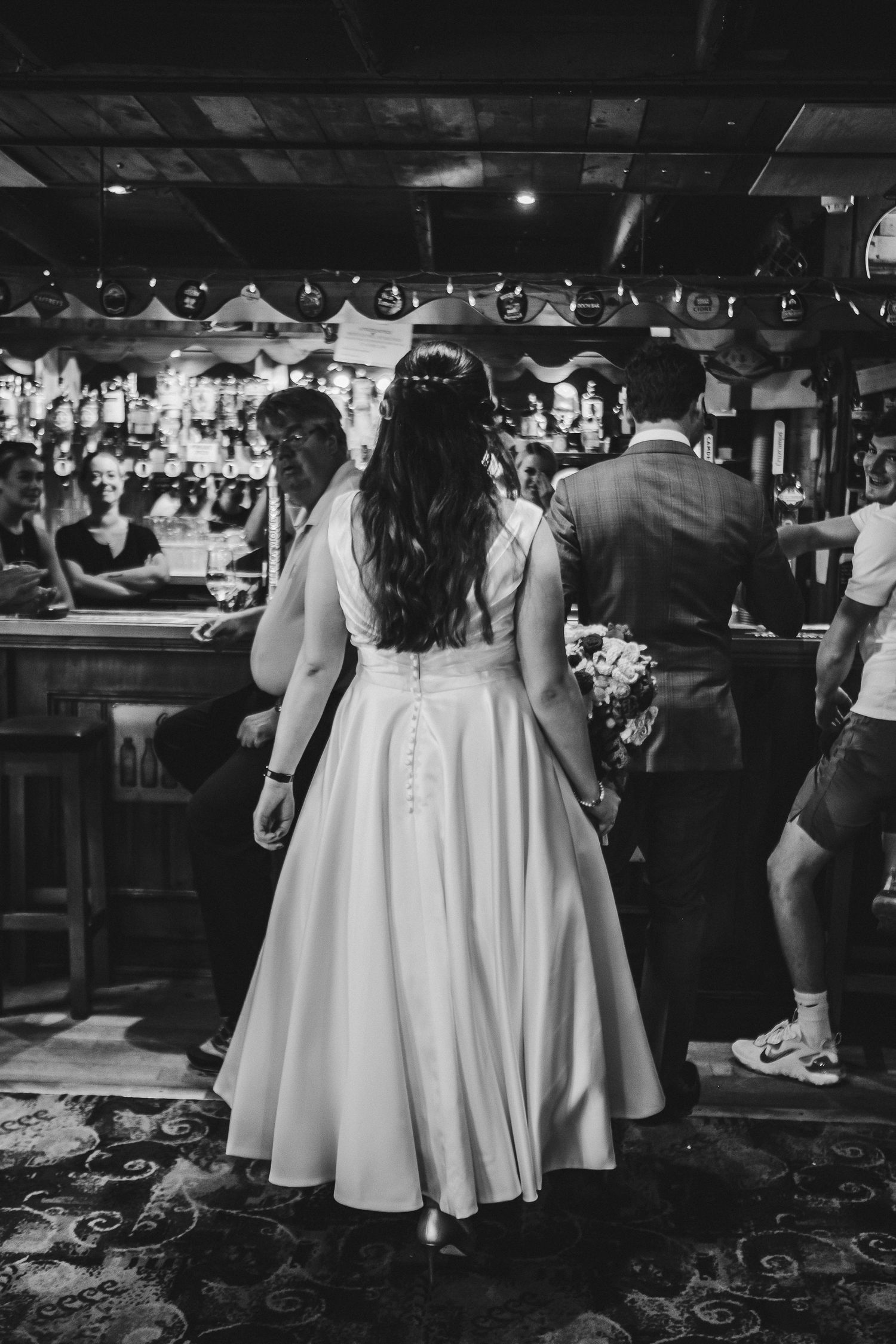 Back view of bride in tea-length dress at bar with wedding reception guests.