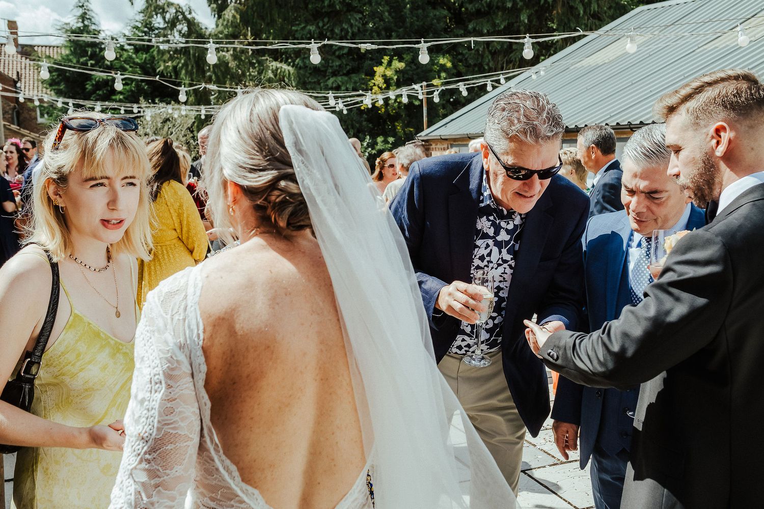 Wedding guests gather around during an outdoor celebration under a string-lit canopy.