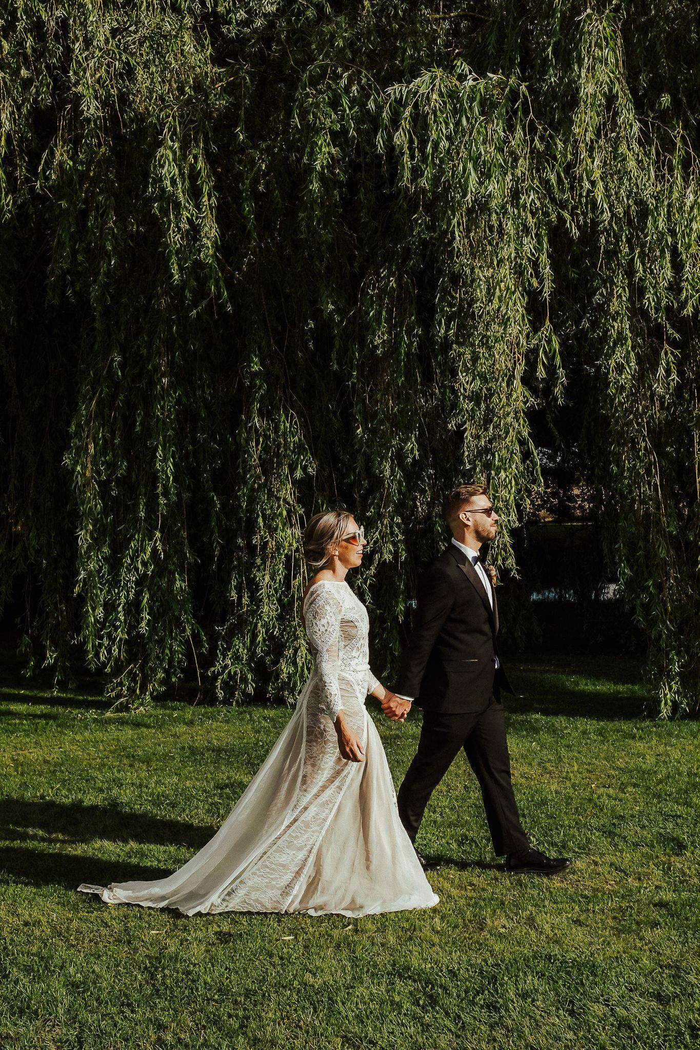 Wedding couple walking under weeping willow trees in formal attire.