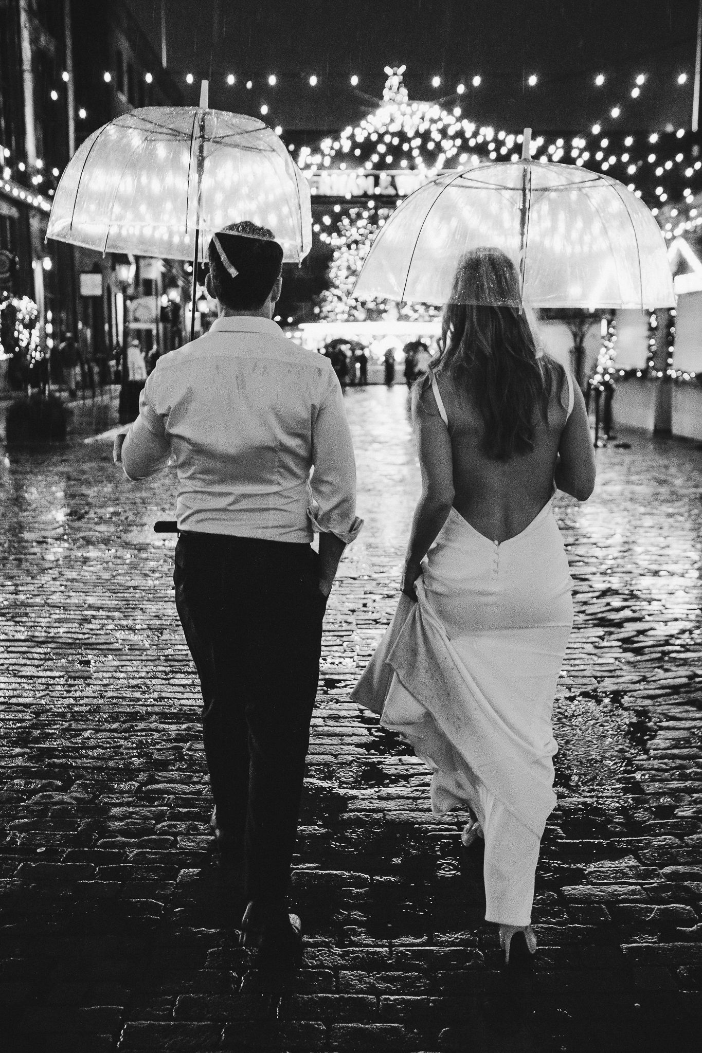 Couple with clear umbrellas walking on wet cobblestone street under holiday lights.