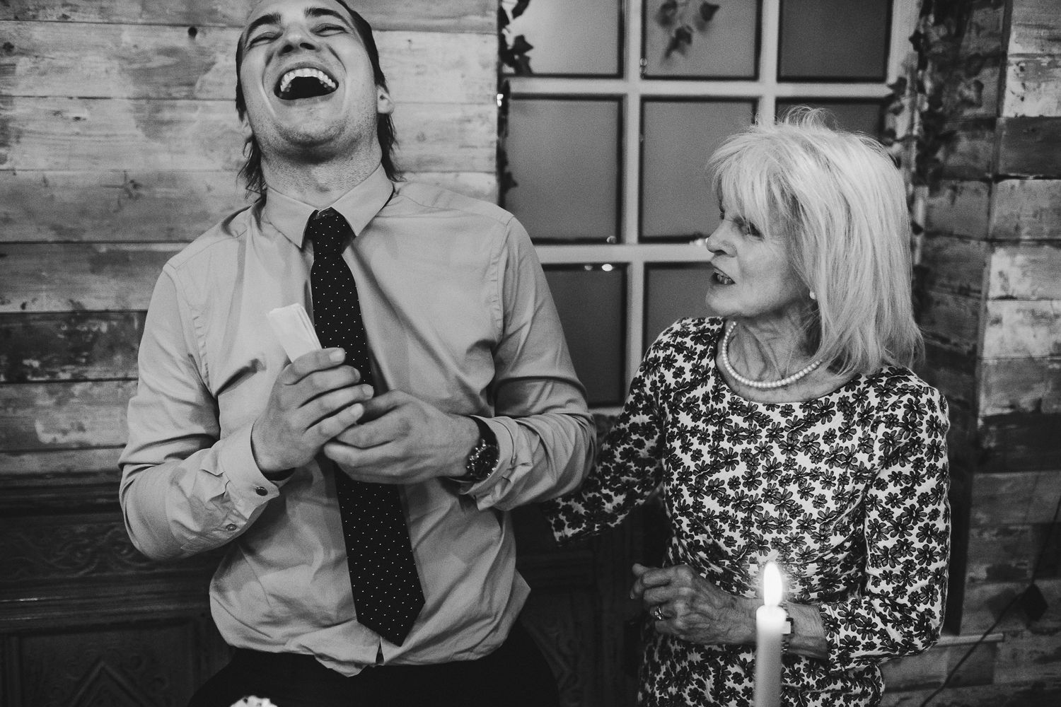 Two people share a candid moment of laughter by candlelight against a rustic wooden wall.