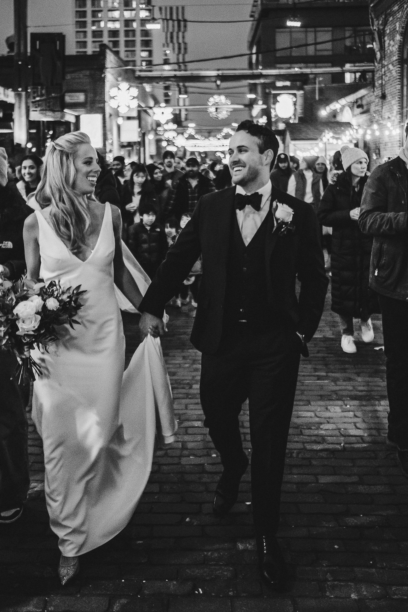 Couple walks through city streets at night with festive lights overhead.