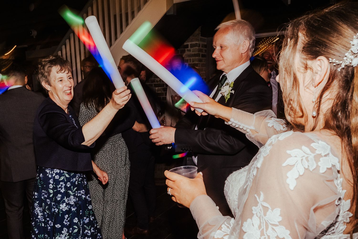 Wedding reception guests dance with glowing LED batons in a dimly lit venue.