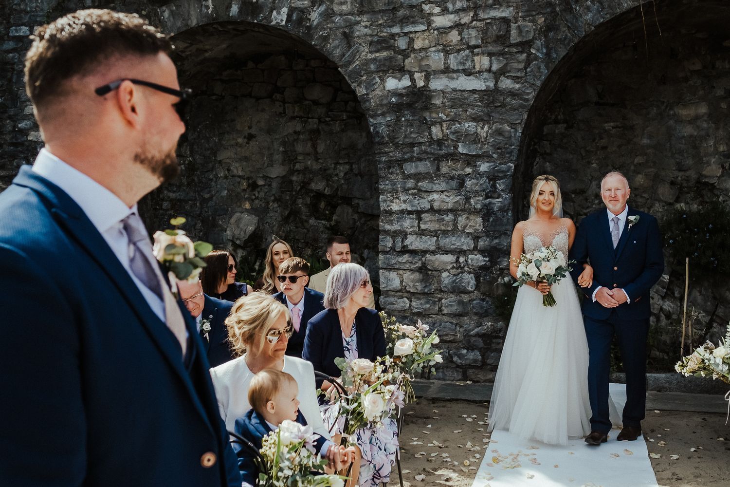 A wedding ceremony takes place under stone archways with floral decorations and gathered guests.