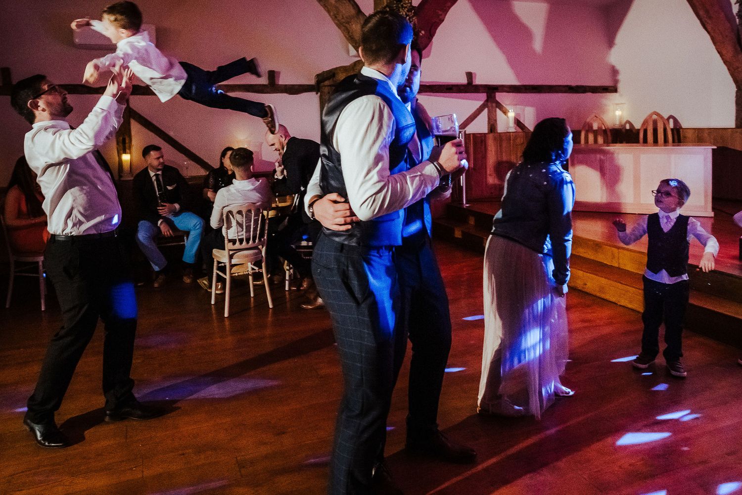 Wedding guests dancing energetically on a wooden dance floor with colorful lighting in the background.