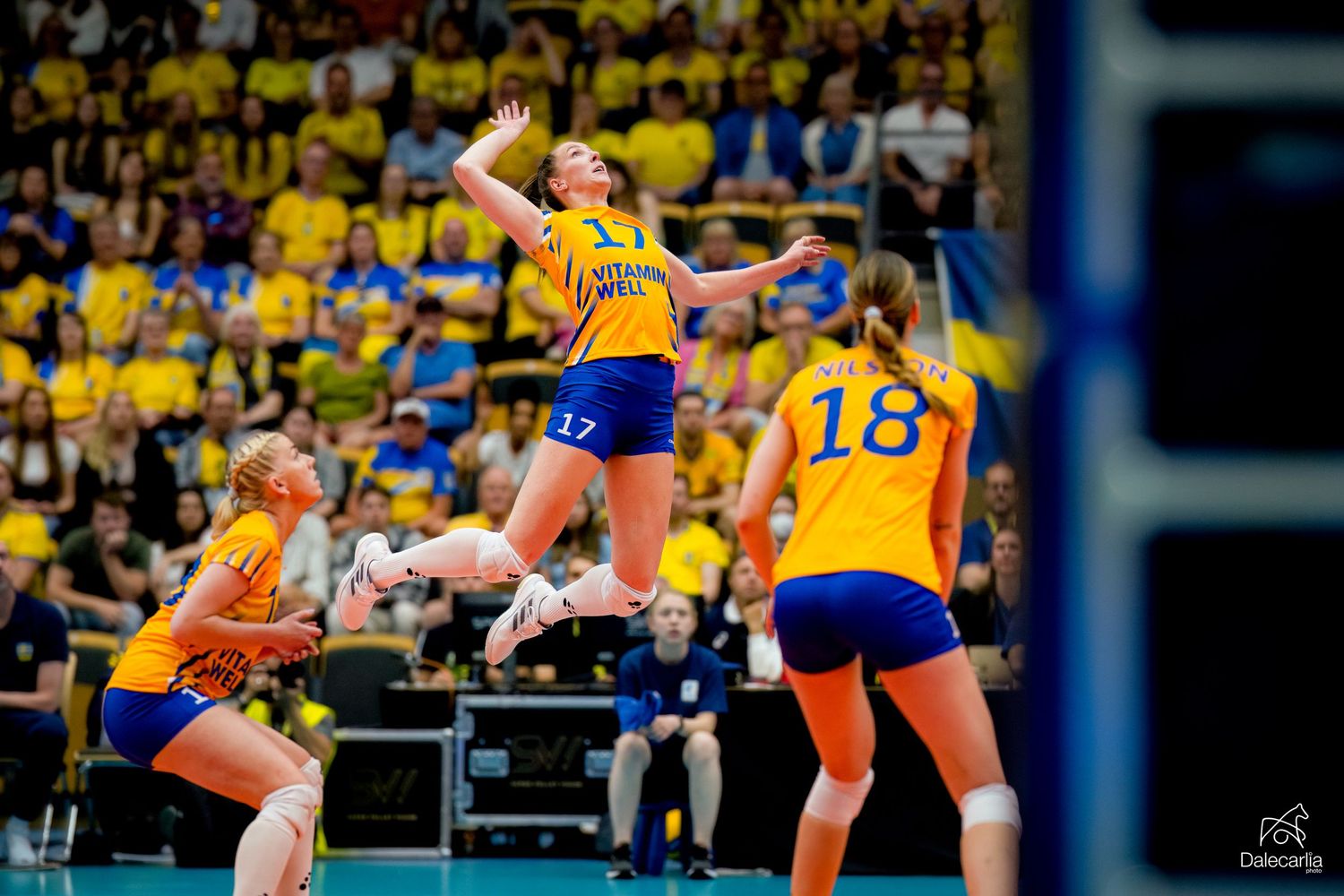 Volleyball players in yellow and orange uniforms mid-action during an intense match with a leaping player in focus.