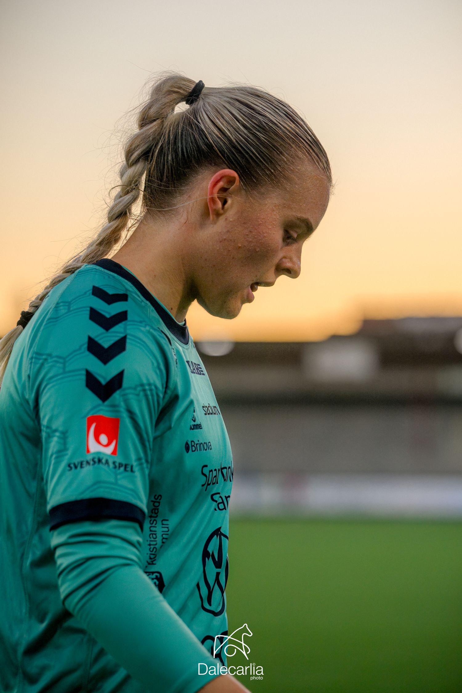 Soccer goalkeeper in turquoise jersey looking down thoughtfully during sunset at a sports field.