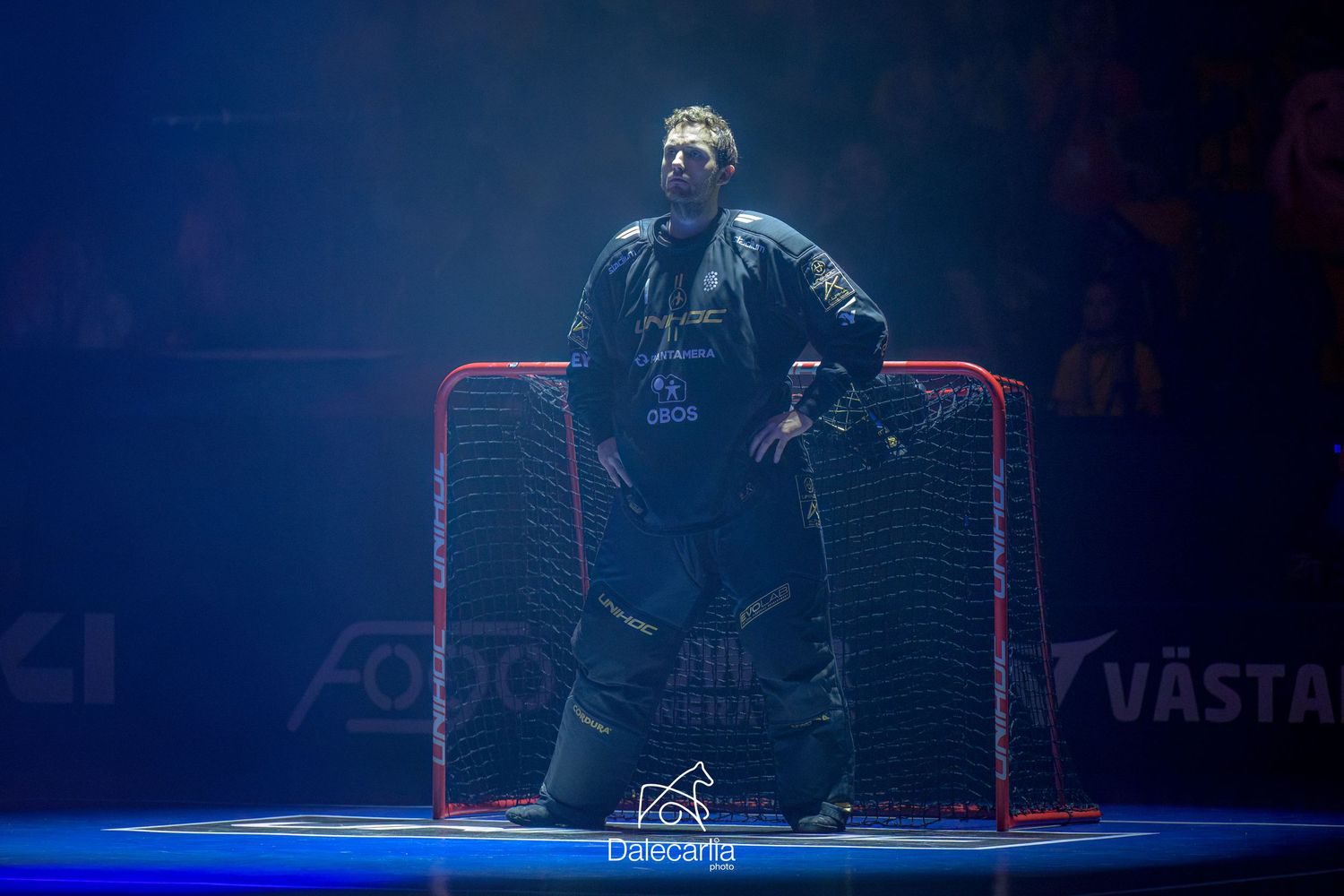 Ice hockey goalie in protective gear stands in front of a red goal net illuminated by blue lighting during a game.