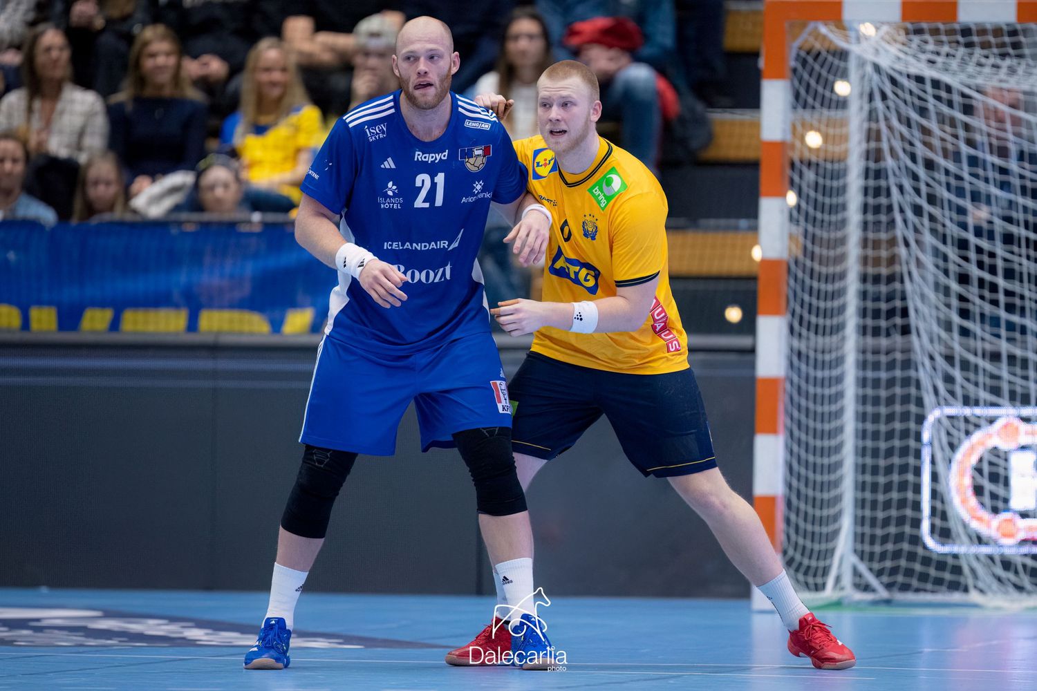 Indoor handball match action shows players from blue and yellow teams battling near the goal post during competitive play.