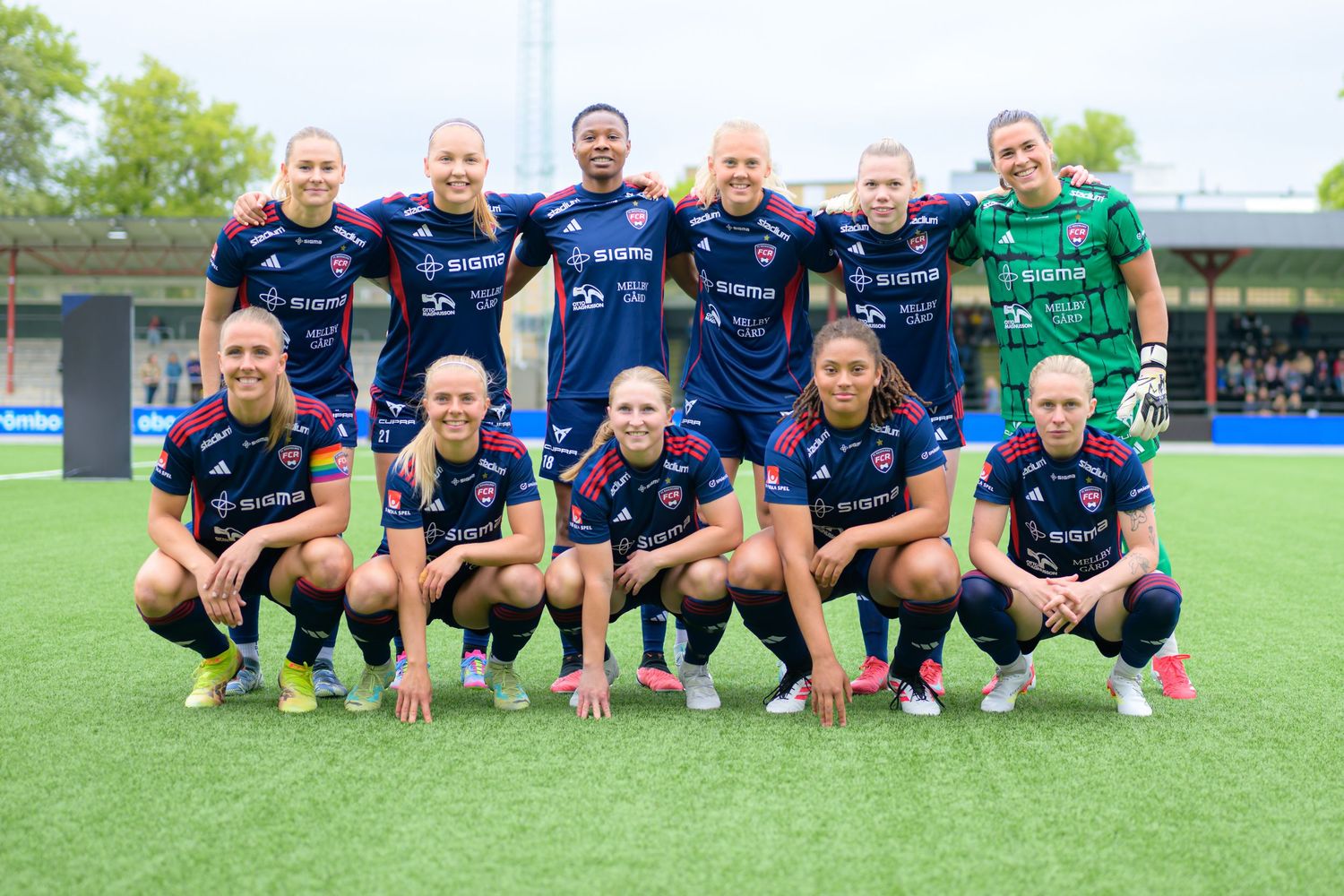 Soccer team poses for group photo on artificial turf field wearing navy blue and green uniforms.