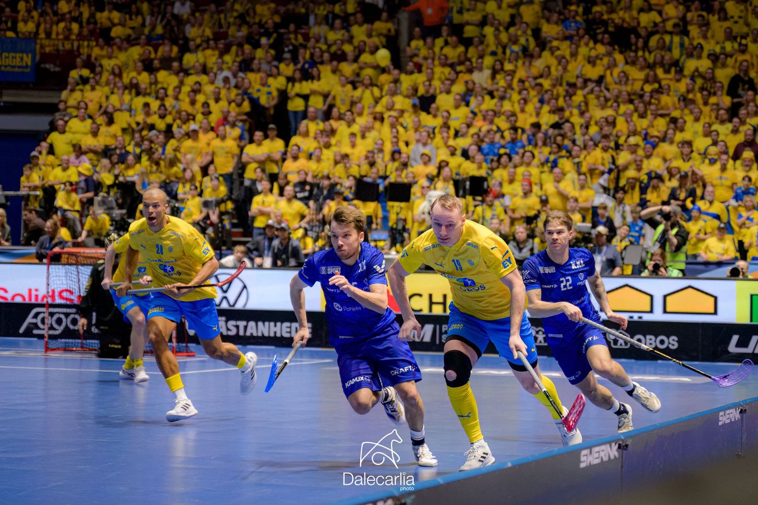 Professional handball players in blue and yellow uniforms compete in an intense match in front of packed yellow crowd.