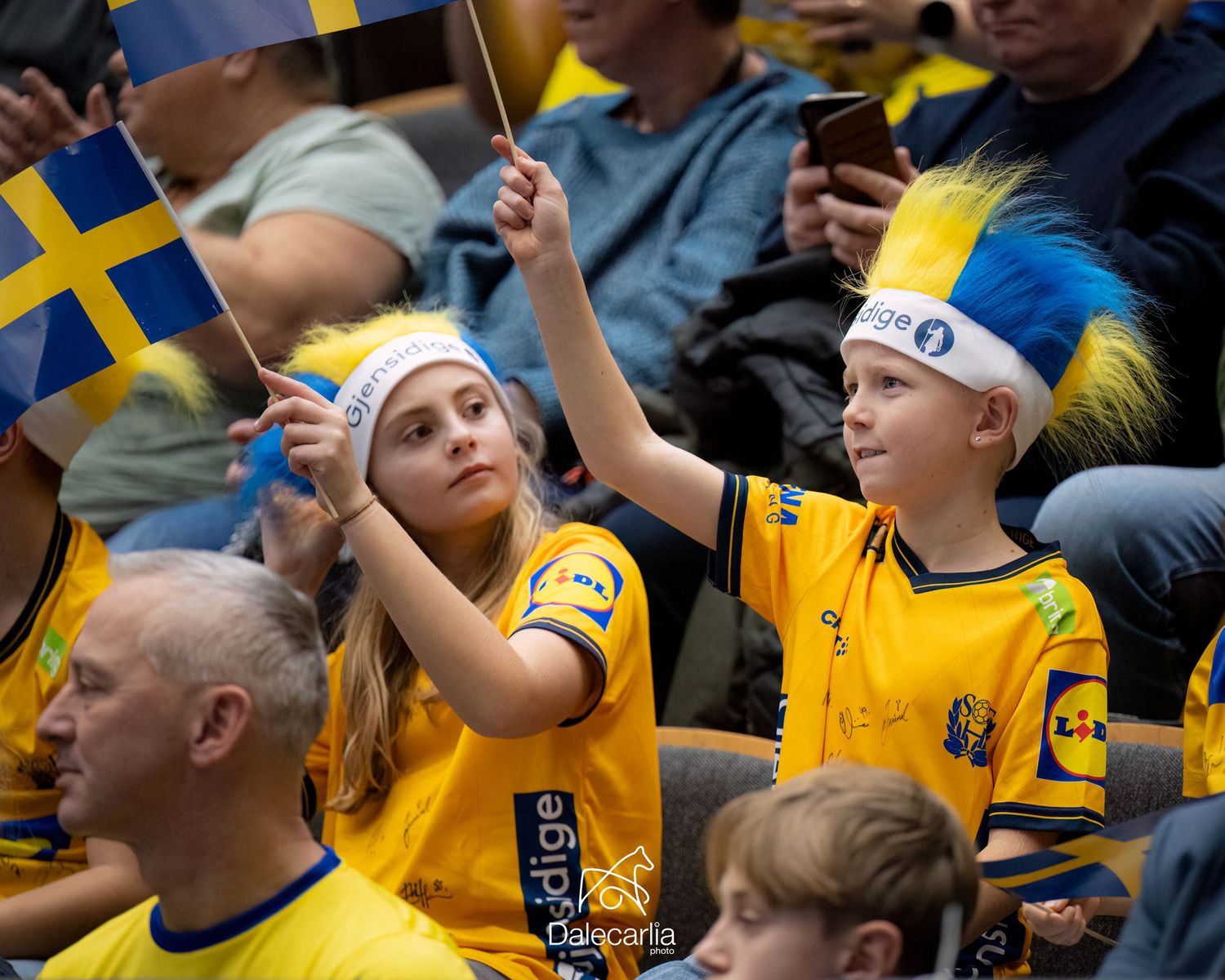 Young Swedish soccer fans wearing yellow jerseys and team headbands cheer during a match at the stadium.