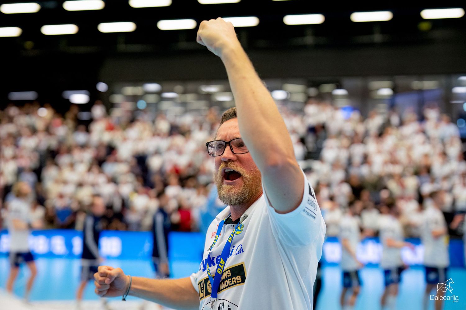 Coach celebrates with raised fist and passionate expression during indoor sporting event with crowd in background.
