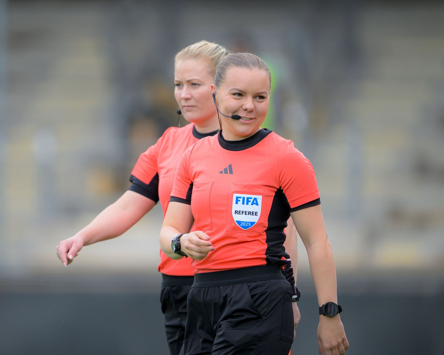 Two soccer referees in bright coral colored jerseys and black shorts walking on a sports field during a match.