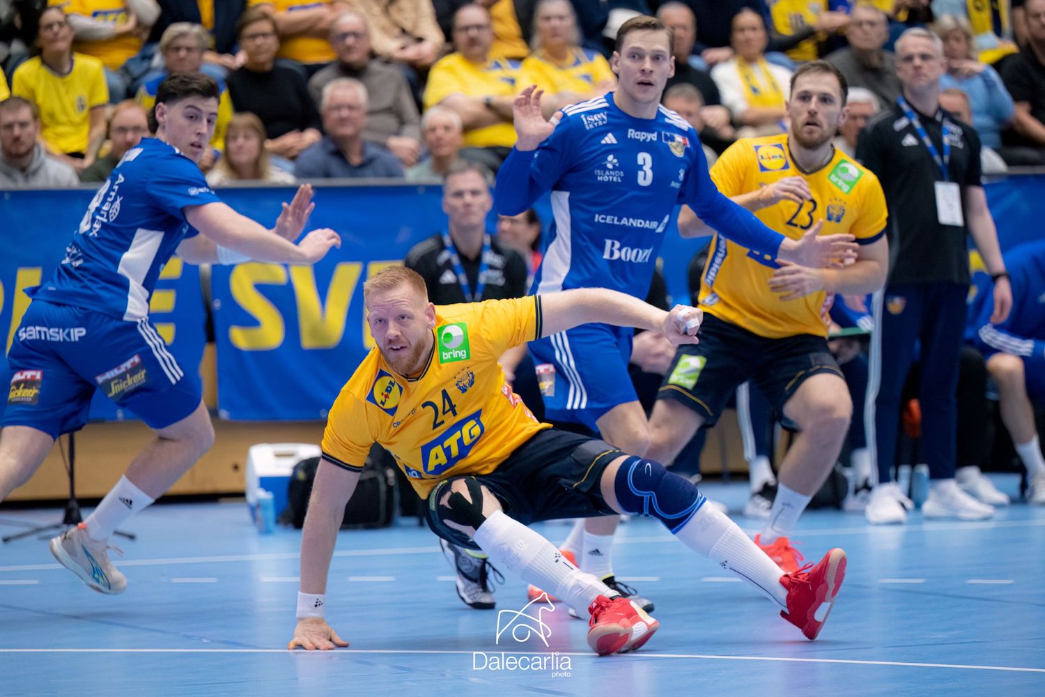 Indoor handball match action shows players in yellow and blue uniforms colliding during an intense play on the court.