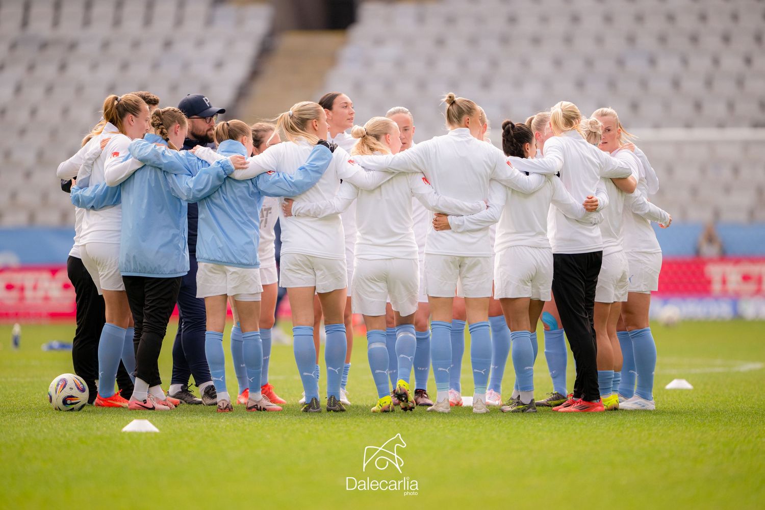 Soccer team huddles together in a circle on the field wearing light blue and white uniforms before a match.