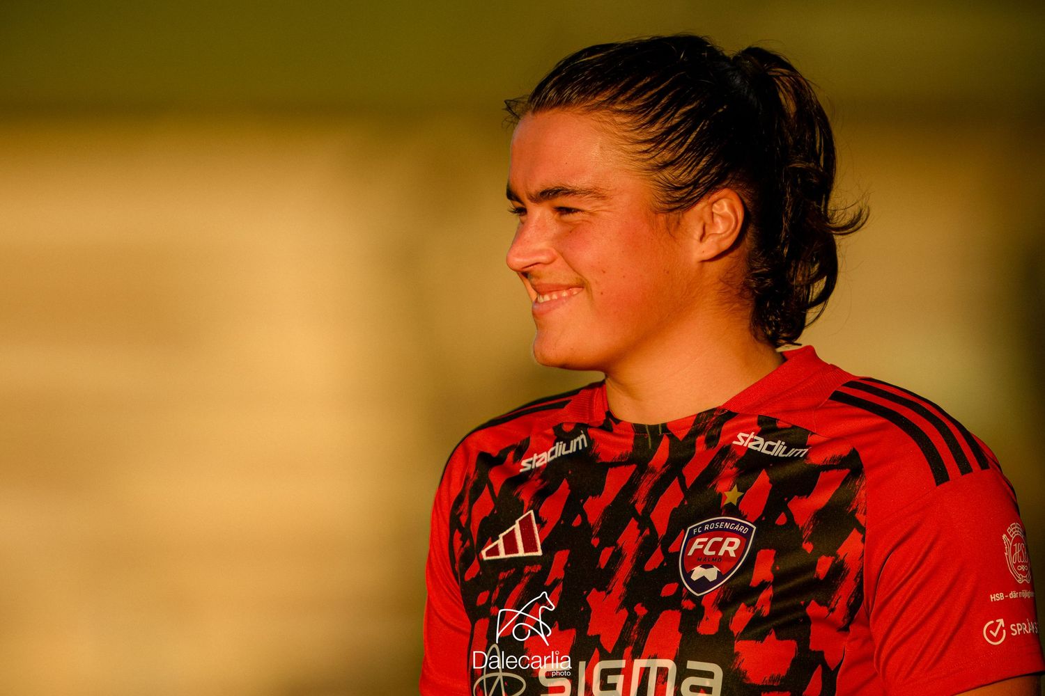 A soccer player in a red Adidas jersey looks to the side while bathed in warm golden sunlight.