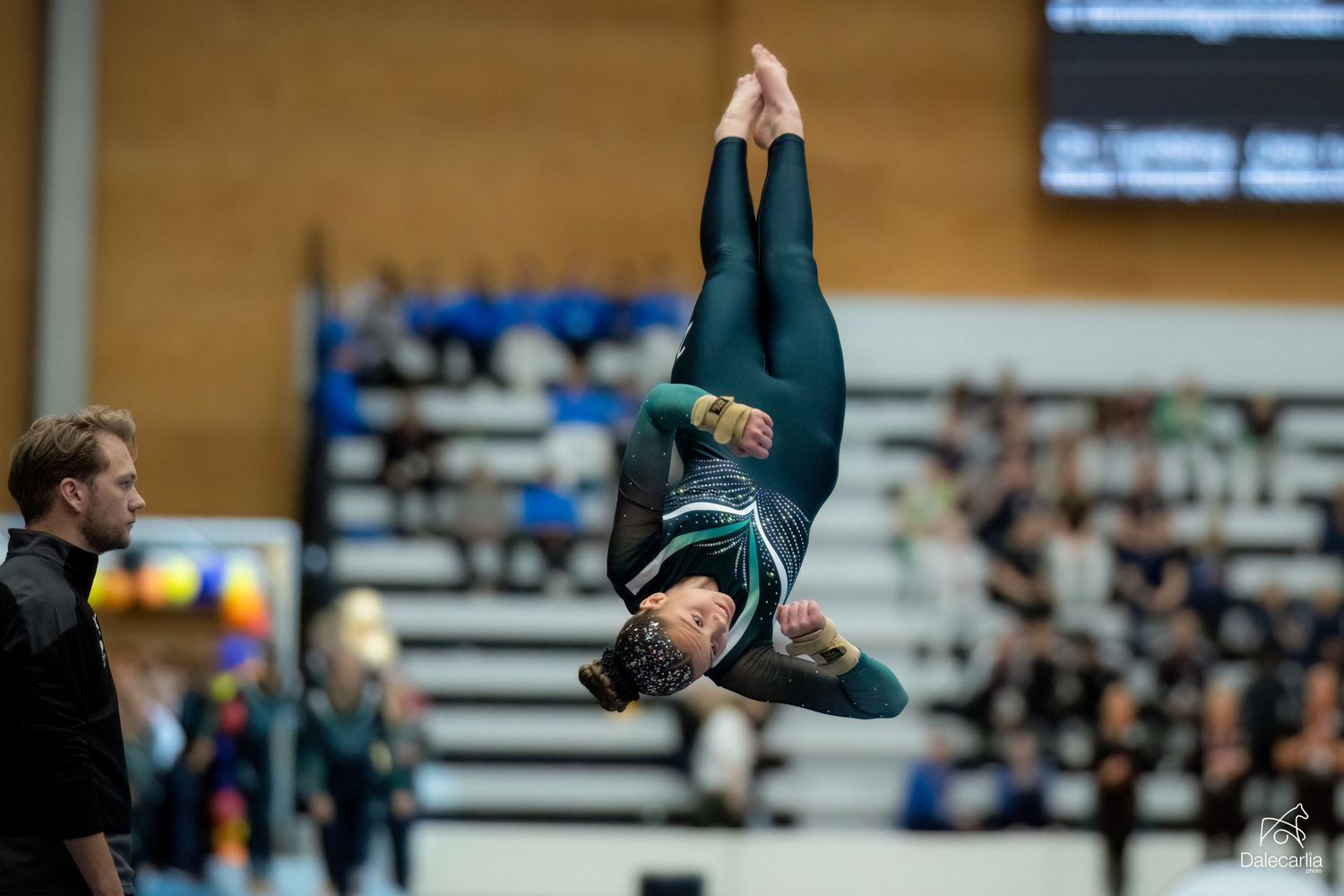 A gymnast performs an inverted aerial maneuver during a competition in a gymnasium with spectators in the background.