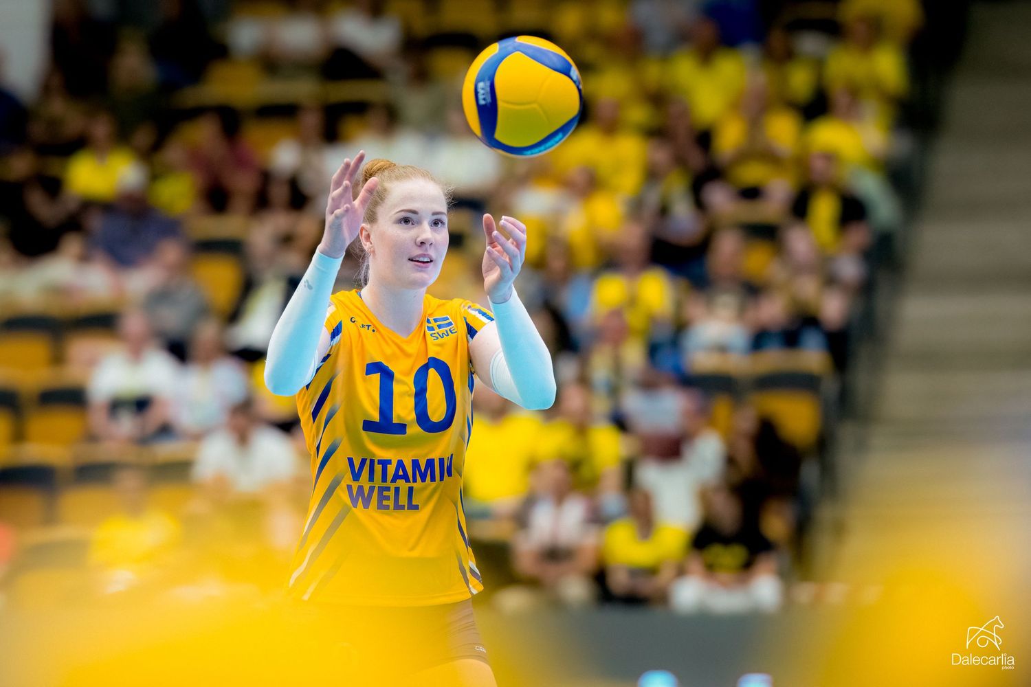 Female volleyball player in yellow jersey number 10 watches the ball during an intense match with crowd in background.
