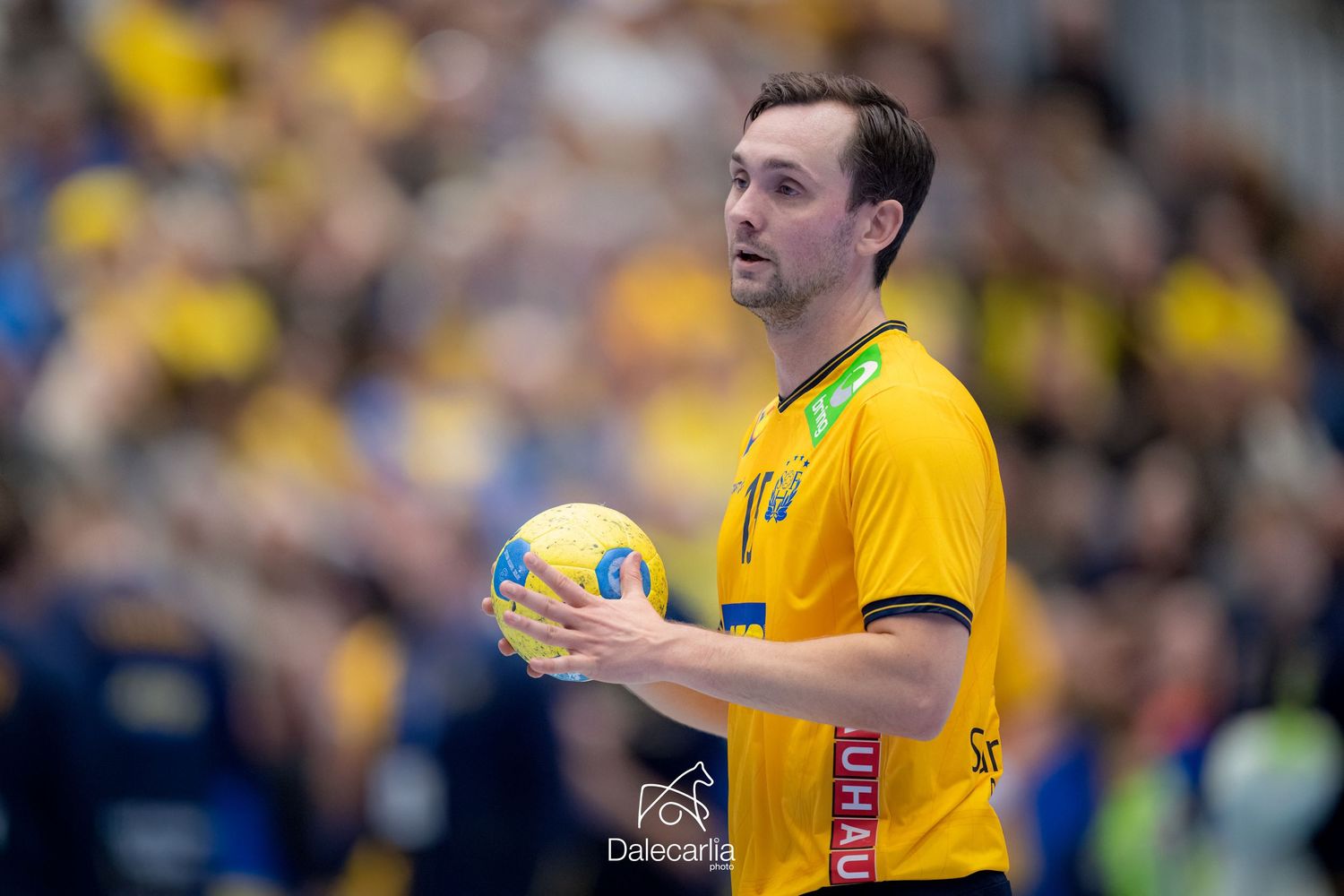 Handball player in yellow uniform holds ball during competitive match with spectators in background.