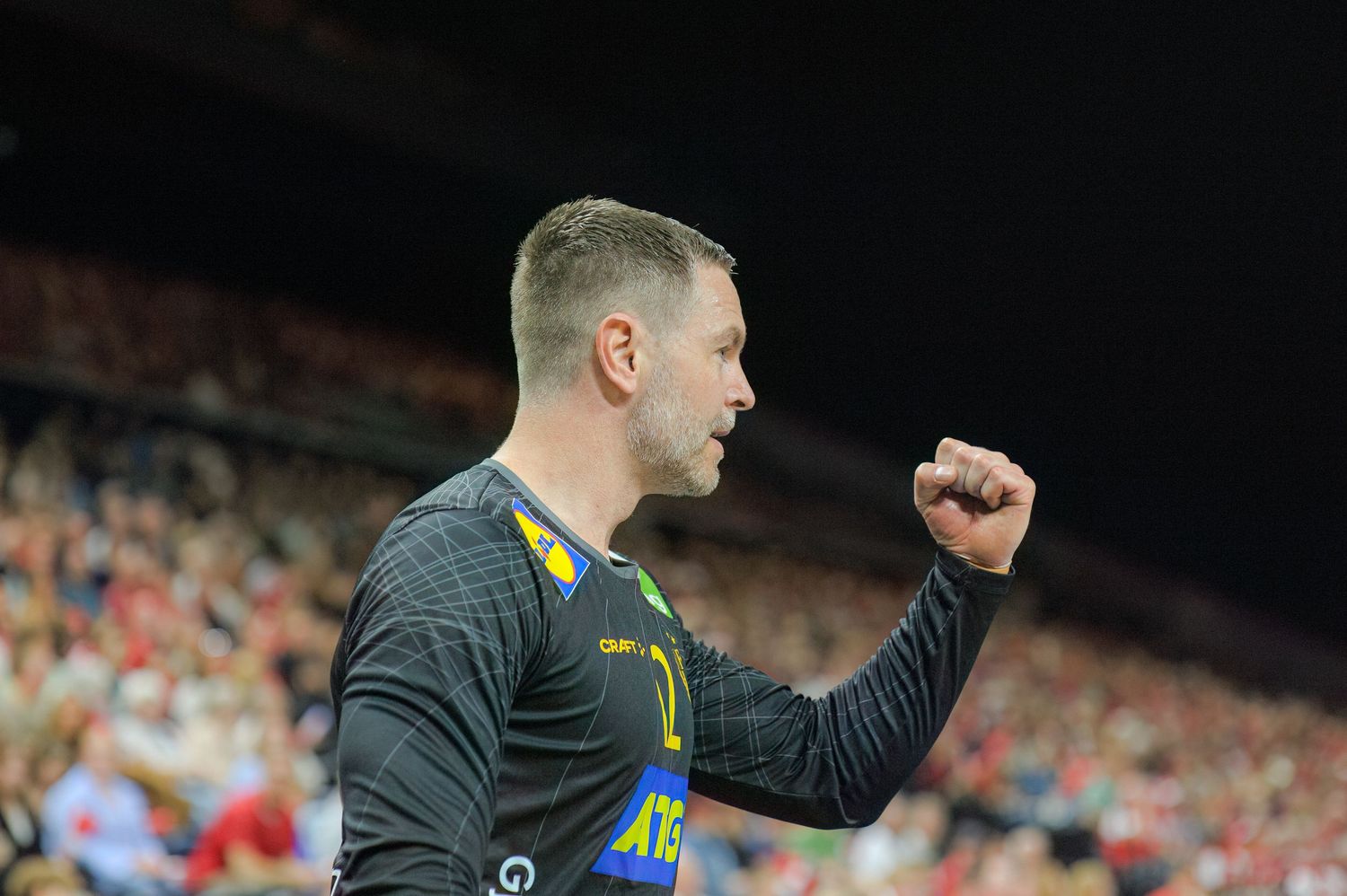 A soccer player wearing a dark grey jersey celebrates with a raised fist during a match in a stadium.