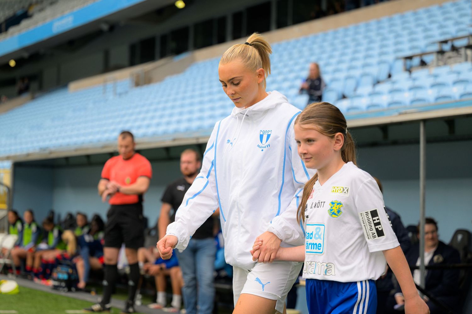 A player in white uniform walks onto a soccer field with stadium seating and blue chairs visible in the background.