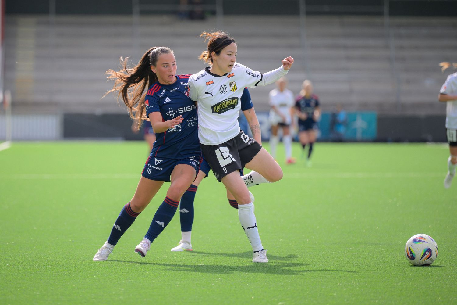 Two soccer players from opposing teams battle for possession of the ball during a competitive match on an artificial turf field.