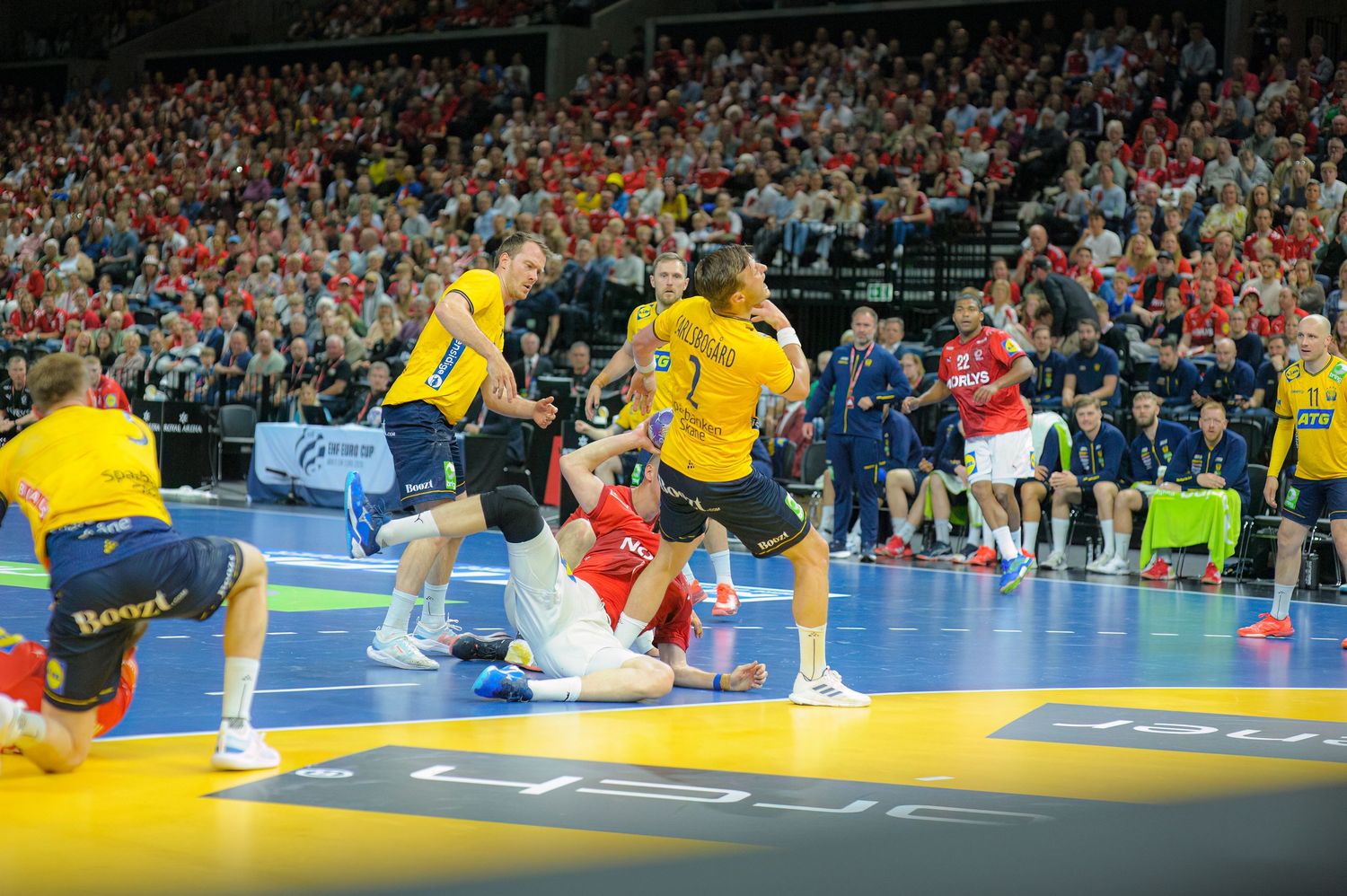 Handball players in yellow and red uniforms compete intensely during a professional match on a blue court.